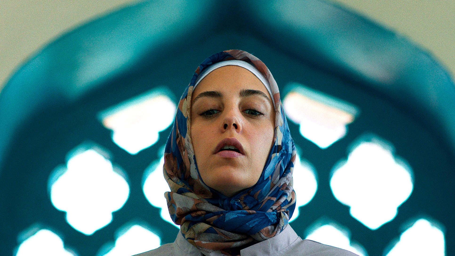 L'image montre une femme portant un hijab coloré, se tenant dans un cadre architectural. La lumière met en valeur son visage et le fond présente des motifs géométriques typiques des designs islamiques, avec des arches et des vitraux. Son expression est sérieuse, ce qui ajoute à la profondeur de la scène.