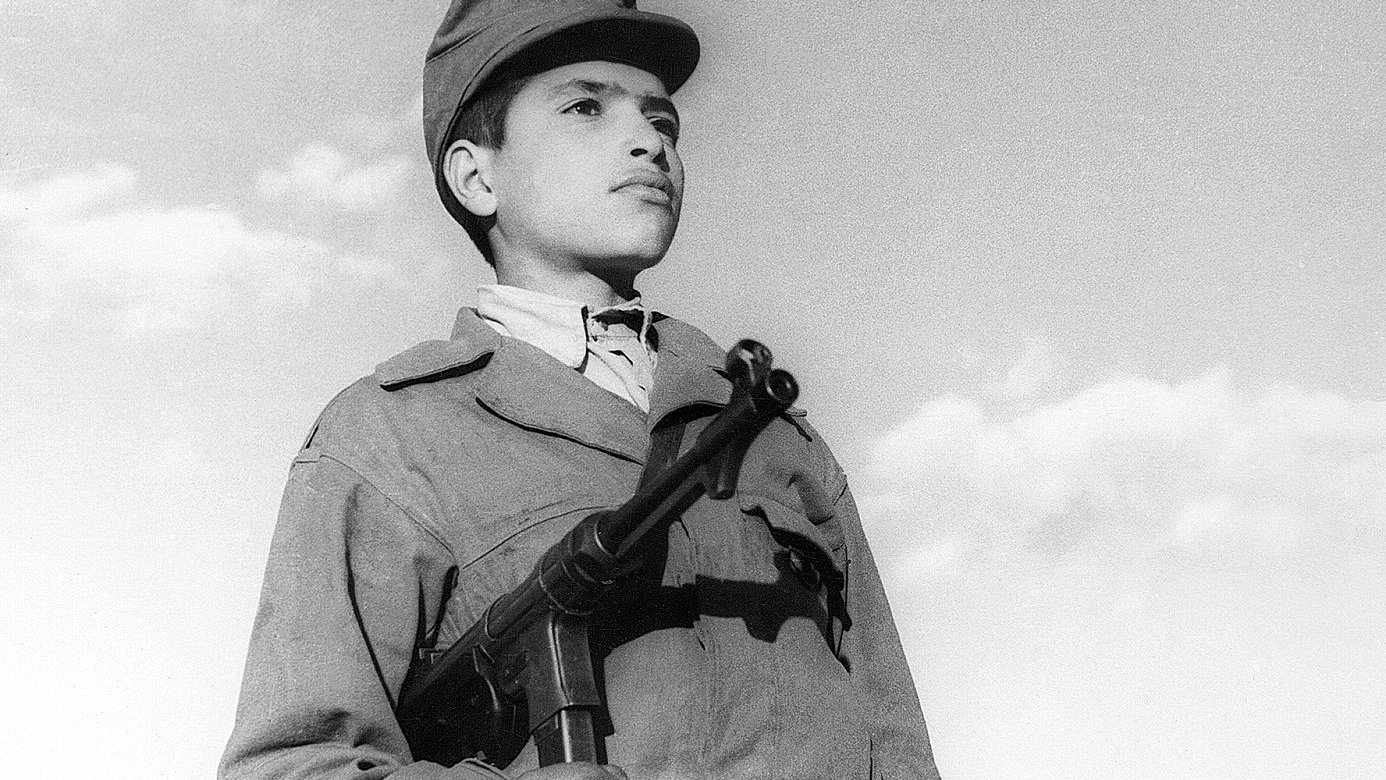 L'image montre un jeune garçon en uniforme militaire, tenant une arme à feu. Son regard est sérieux et déterminé, tandis qu'il se tient droit, face à l'objectif. Le ciel en arrière-plan est clair, avec quelques nuages. L'uniforme et l'arme peuvent indiquer qu'il fait partie d'un contexte militaire ou historique spécifique. L'ambiance de la photo évoque un sentiment de courage et de bravoure.