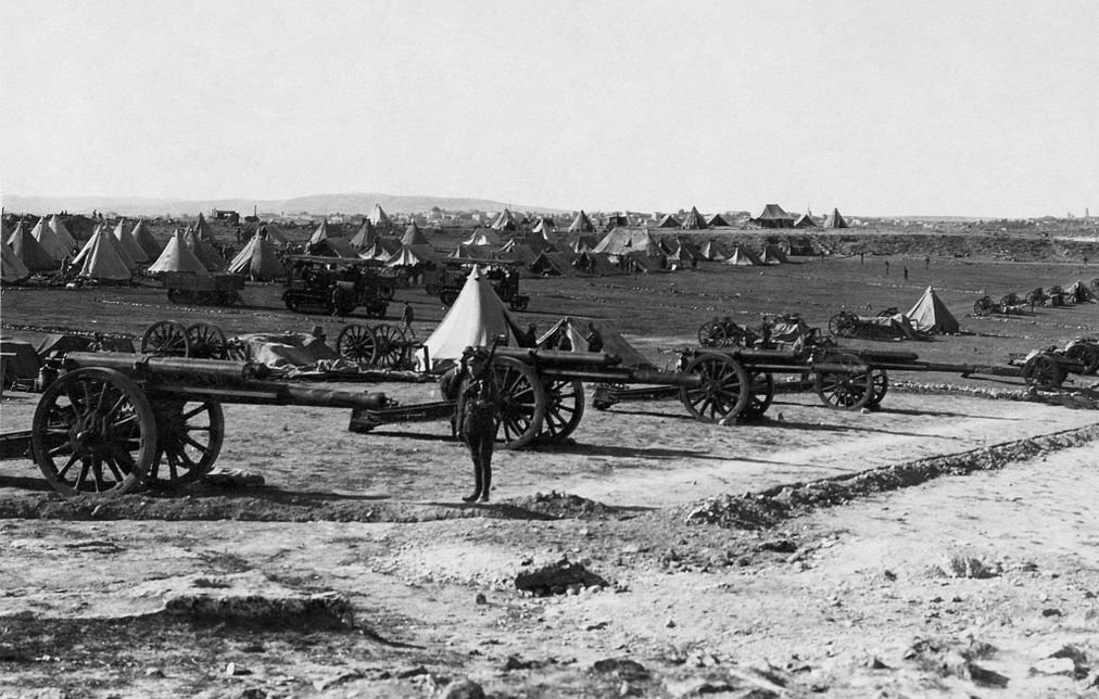 L'image montre un camp militaire avec plusieurs canons en premier plan. À l'arrière-plan, on distingue des tentes disposées de manière ordonnée sur un terrain dégagé. Le paysage semble vaste, avec des collines ou des montagnes au loin. L'atmosphère est historique, évoquant une période de guerre ou de mobilisation des troupes. Un soldat ou un homme se tient près des canons, donnant une échelle à la scène.