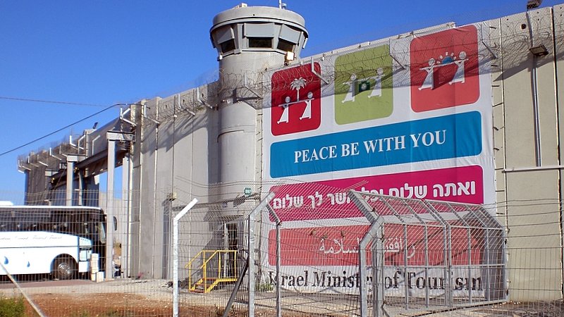 L'image montre un mur imposant, probablement un mur de séparation, avec une tour de surveillance au sommet. À la base du mur, il y a une grande pancarte qui affiche des messages liés à la paix, écrits en plusieurs langues. L'environnement est urbain, et on peut voir un bus stationné à proximité. Le tout est entouré d'une clôture barbelée, ce qui donne une impression de sécurité renforcée. Le ciel est dégagé et ensoleillé.