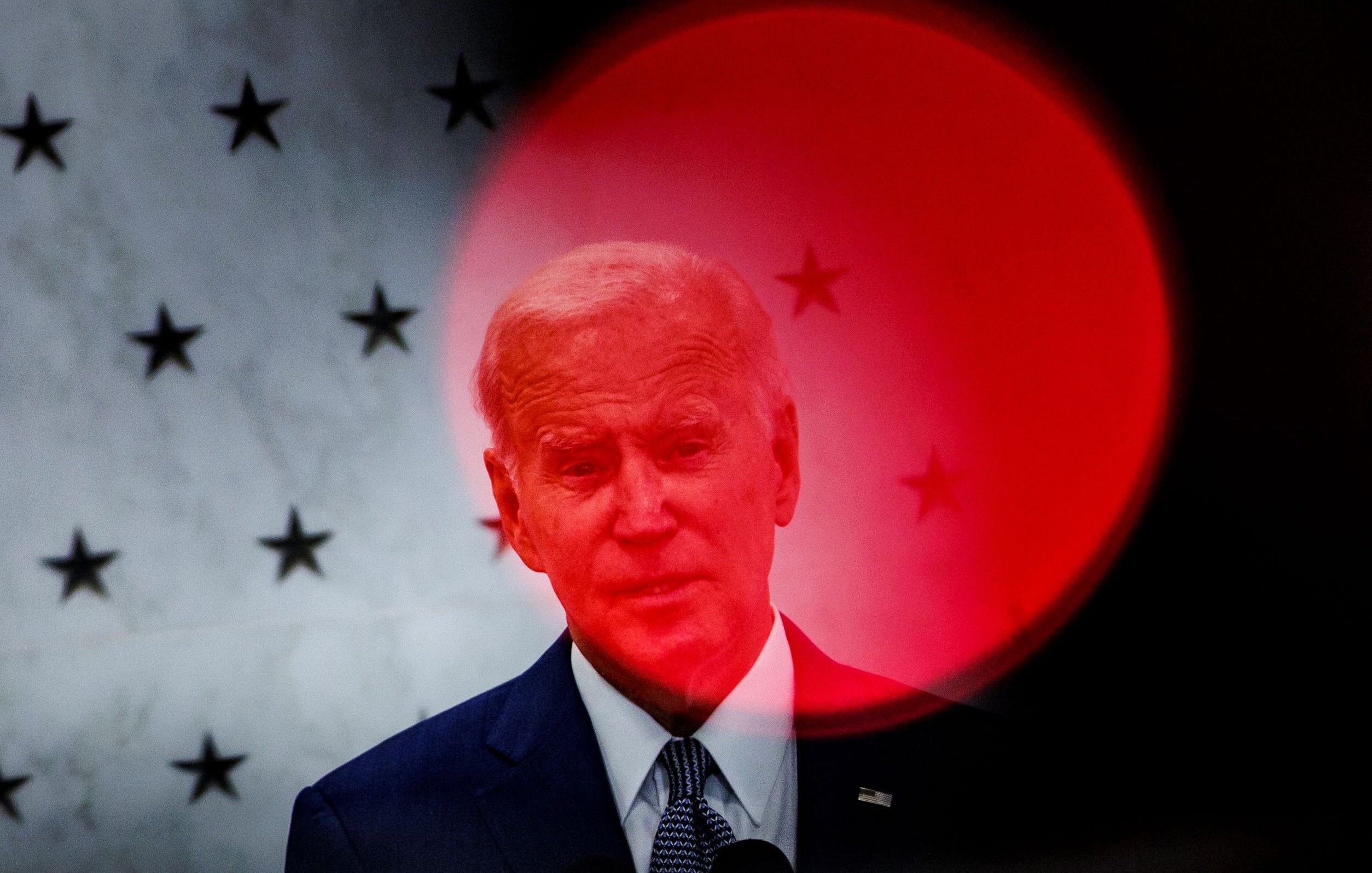 L'image montre un homme en train de parler, probablement lors d'un discours. En arrière-plan, on peut voir un motif de petites étoiles qui s'étale sur une surface claire. Une lumière rouge qui semble provenir de l'avant-plan crée un flou, accentuant la figure de l'homme et lui donnant une apparence distincte. L'atmosphère de l'image semble sérieuse ou solennelle, suggérant un événement important.