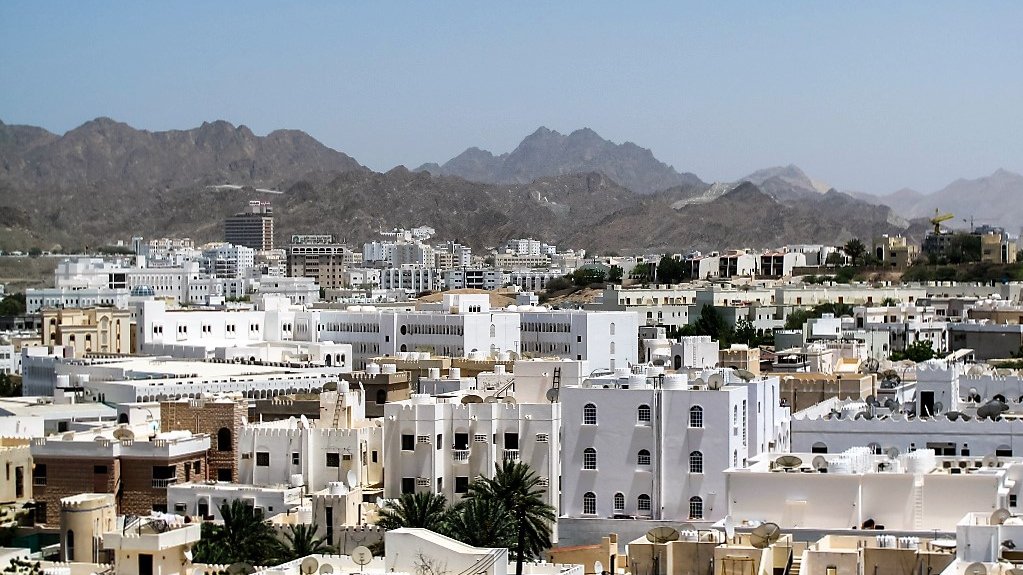 L'image montre une vue panoramique d'une ville située dans une région montagneuse. On peut voir de nombreux bâtiments aux façades blanches et crème, qui typiquement se distinguent dans un environnement aride. Les montagnes en arrière-plan ajoutent une dimension naturelle impressionnante au paysage urbain. Le ciel est dégagé et ensoleillé, accentuant la lumière qui se reflète sur les bâtiments. Cette scène évoque une atmosphère calme et sereine.