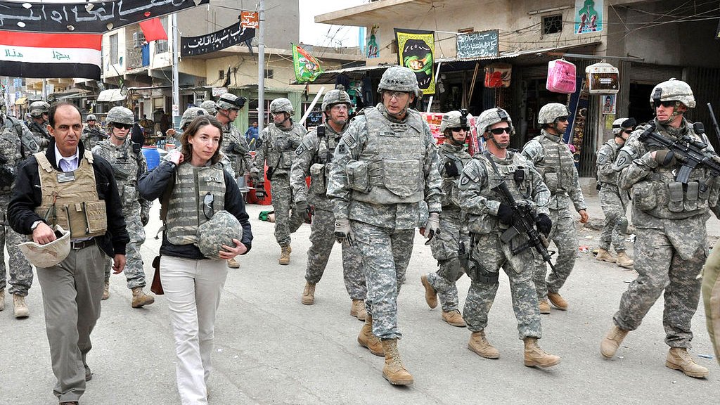 L'image montre un groupe de soldats en uniforme militaire marchant dans une rue. Ils semblent être dans une zone urbaine, possiblement dans un pays en conflit. Au premier plan, on voit une femme en vêtements civils, portant un gilet pare-balles, marchant à côté d'un homme également en tenue civile. Le décor autour d'eux comprend des bâtiments, des panneaux et d'autres signes de vie urbaine. L'atmosphère semble sérieuse, reflétant un environnement potentiellement tendu.