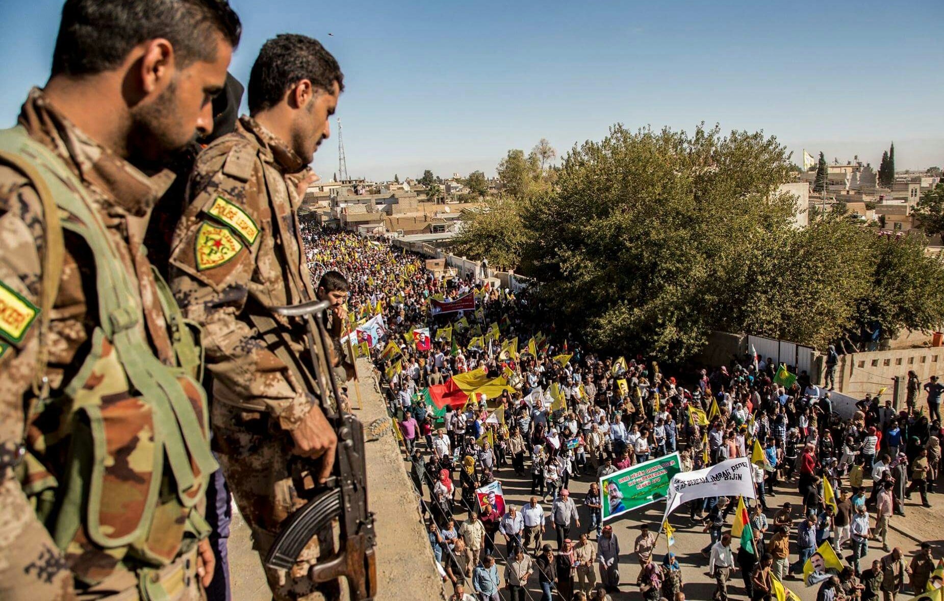L'image montre une scène de manifestation en plein air, où une grande foule est rassemblée, tenant des drapeaux et des pancartes. Au premier plan, deux soldats armés, portant des uniformes camouflés, surveillent la foule depuis un point élevé. On peut voir des arbres et des bâtiments en arrière-plan, et le ciel est dégagé. L'atmosphère semble chargée d'émotion, avec des participants visiblement engagés.