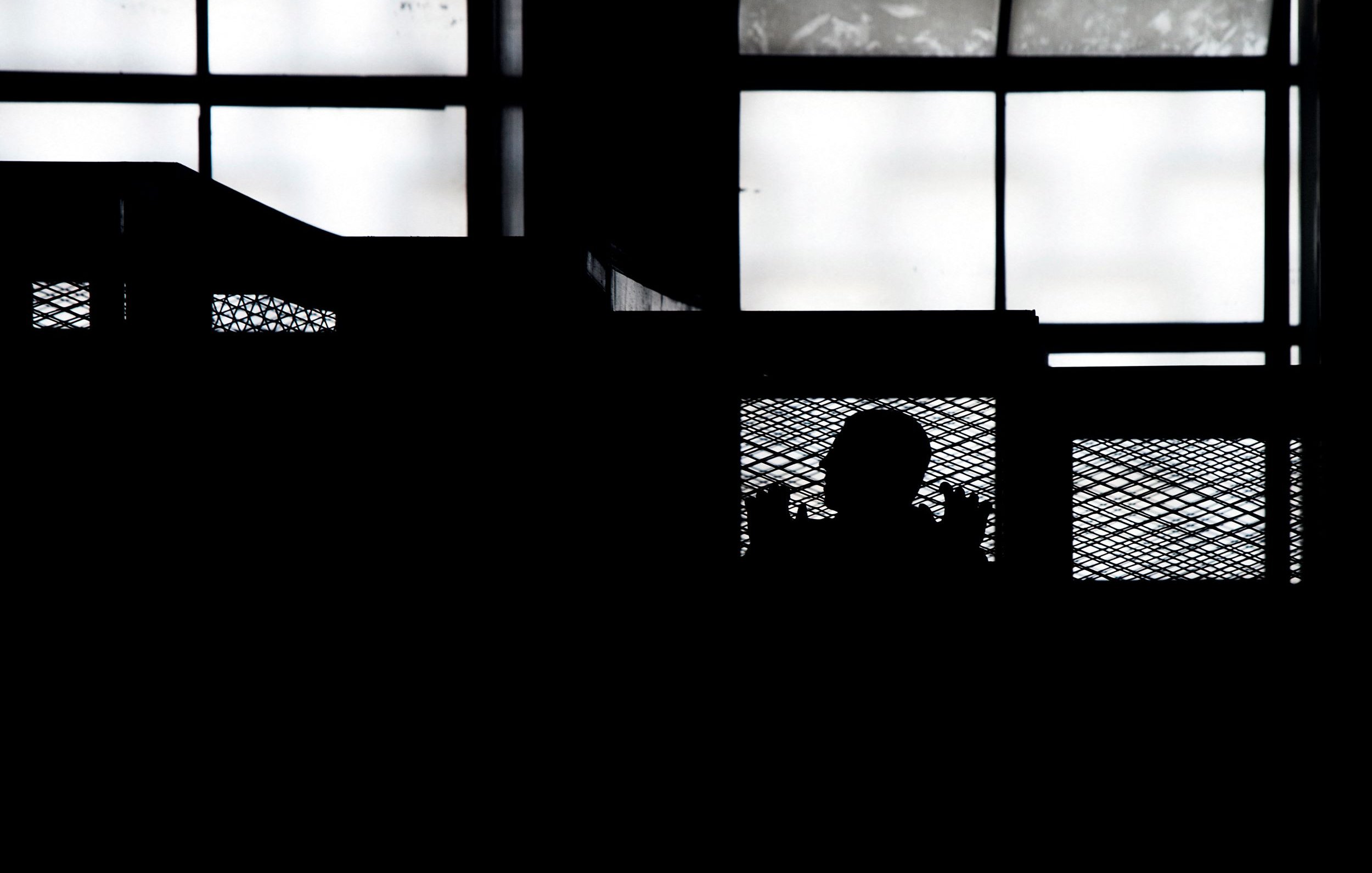 L'image montre une silhouette, probablement d'une personne, derrière une structure en métal qui semble être une grille ou une barrière. En arrière-plan, on aperçoit des fenêtres laissant passer une lumière diffuse. L'ensemble de la scène est dominé par des ombres, créant une atmosphère mystérieuse. Les contours des objets sont flous, ce qui accentue l'aspect dramatique de l'image.