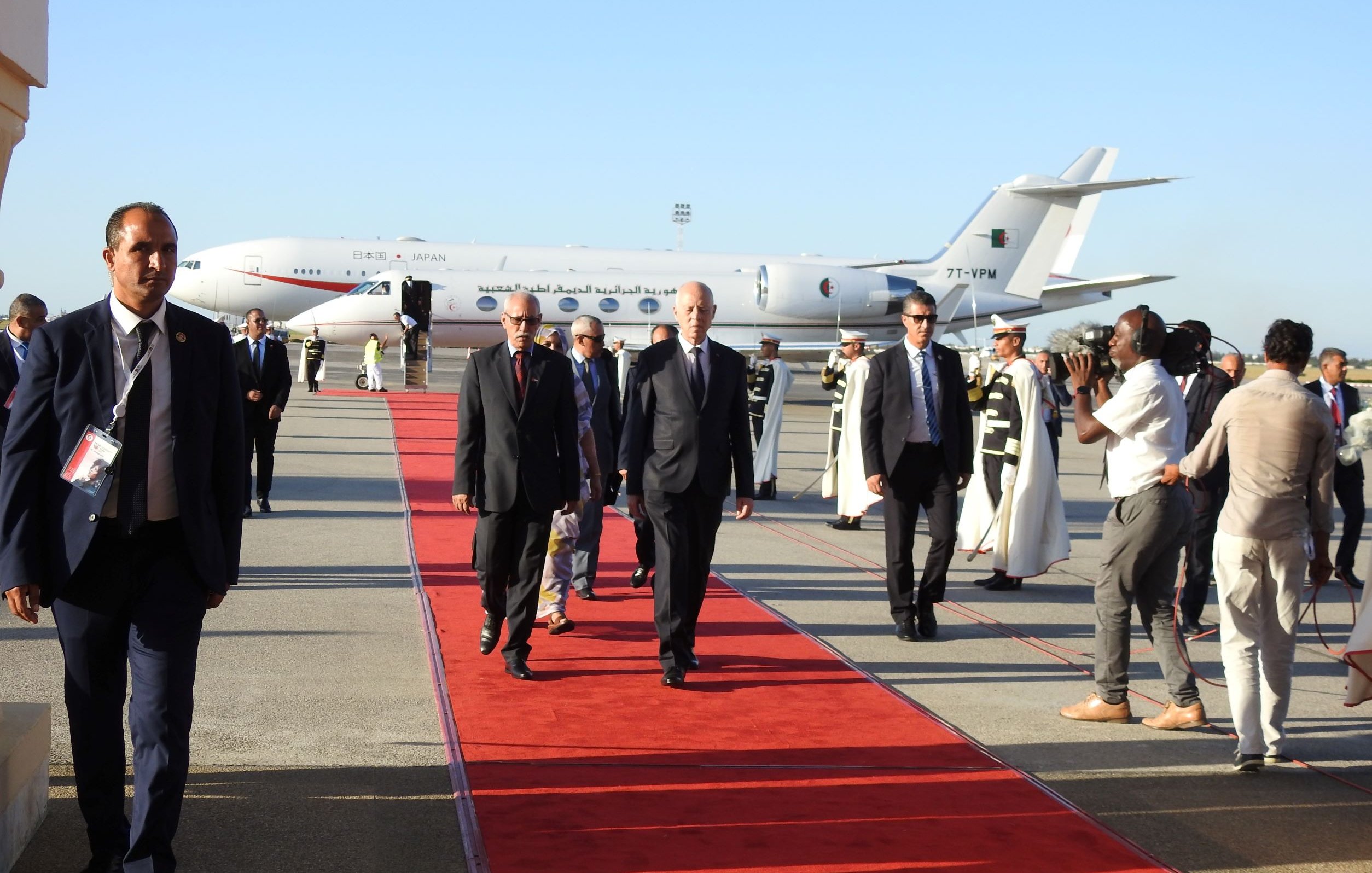 L'image montre un aéroport avec une piste. Un avion, visible en arrière-plan, est stationné. Des personnes marchent sur un tapis rouge, probablement des dignitaires ou des personnalités importantes. Certains hommes en costume accompagnent les personnes sur le tapis, tandis que d'autres, habillés en tenue formelle, semblent filmer ou prendre des photos de l'événement. L'atmosphère est formelle et festive, indiquant une arrivée importante.