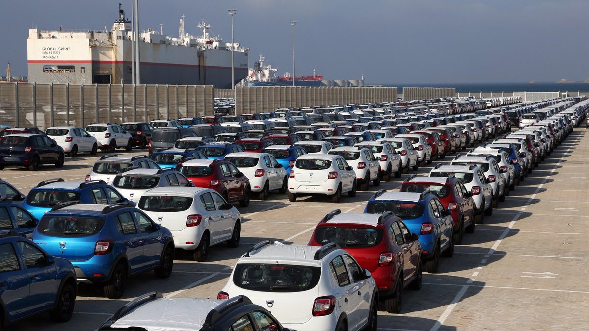 L'image montre un grand parking de voitures, organisé en rangées bien alignées. On aperçoit plusieurs types de voitures de différentes couleurs, majoritairement en blanc, rouge et bleu. En arrière-plan, on peut voir des bateaux de marchandises amarrés dans un port, sous un ciel nuageux. Cette scène semble se dérouler dans une zone portuaire, où les véhicules sont probablement en attente d'expédition ou de distribution.
