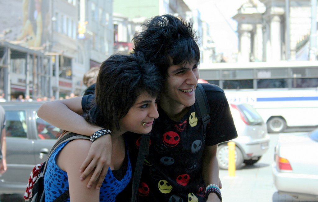 L'image montre deux jeunes personnes souriantes, posant côte à côte dans une rue animée. L'un des jeunes porte un t-shirt noir à motifs colorés, tandis que l'autre a un haut à motifs bleus. Ils semblent heureux et proches, et l'ambiance est vivante, avec des voitures et des piétons en arrière-plan. Les bâtiments en arrière-plan ajoutent à l'environnement urbain.