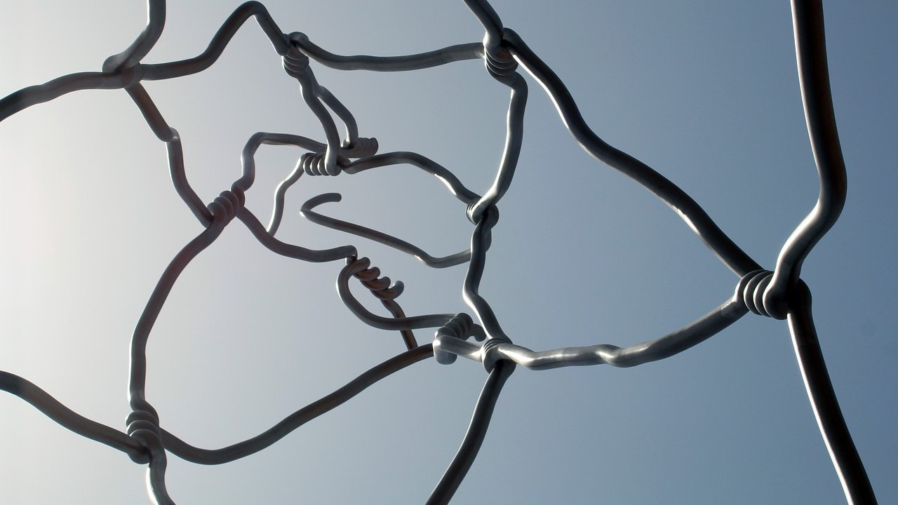 L'image montre une structure artistique composée de fils métalliques entrelacés. Les fils sont courbés et forment des nœuds à certains endroits, créant un motif complexe et abstrait. On peut voir le ciel en arrière-plan, ce qui met en valeur les formes des fils. L'ensemble dégage une impression de légèreté et de fluidité, tout en étant à la fois structurée et organique.