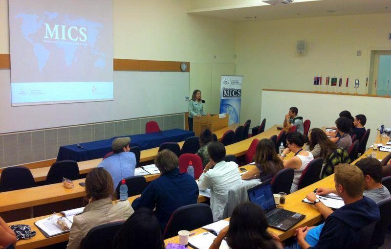 L'image montre une salle de classe ou un auditorium où une présentation est en cours. À l'avant, une intervenante se tient près d'un pupitre, probablement en train de s'adresser à l'auditoire. En arrière-plan, un écran affiche le mot "MICS" accompagné d'une carte du monde. Le public, composé d'une douzaine de personnes, semble attentif et prend des notes sur des ordinateurs portables ou des bloc-notes. La disposition des sièges est en rangées, et quelques personnes sont assises avec des tasses et des boissons. L'ambiance semble académique et interactive.