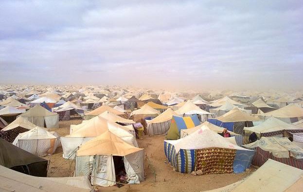 L'image montre un vaste camp de tentes, s'étendant à perte de vue dans un environnement désertique. Les tentes, de diverses tailles et couleurs, sont disposées de manière dense sur le sol sablonneux. Le ciel est nuageux, ce qui donne une atmosphère grise et légèrement menaçante, suggérant peut-être des conditions météorologiques difficiles. On peut également apercevoir de la poussière soulevée par le vent, ajoutant à l'impression de désolation et d'isolement de cet espace.