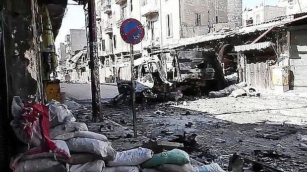 L'image montre une rue dévastée, probablement dans une zone de conflit. On peut voir des bâtiments endommagés et détruits, avec des débris éparpillés sur le sol. Des sacs de sable sont accumulés sur le côté, ce qui suggère des efforts de protection. Un panneau de signalisation est visible, mais l'environnement semble plutôt déserté et en ruines. L'atmosphère dégage un sentiment de désolation et de destruction.