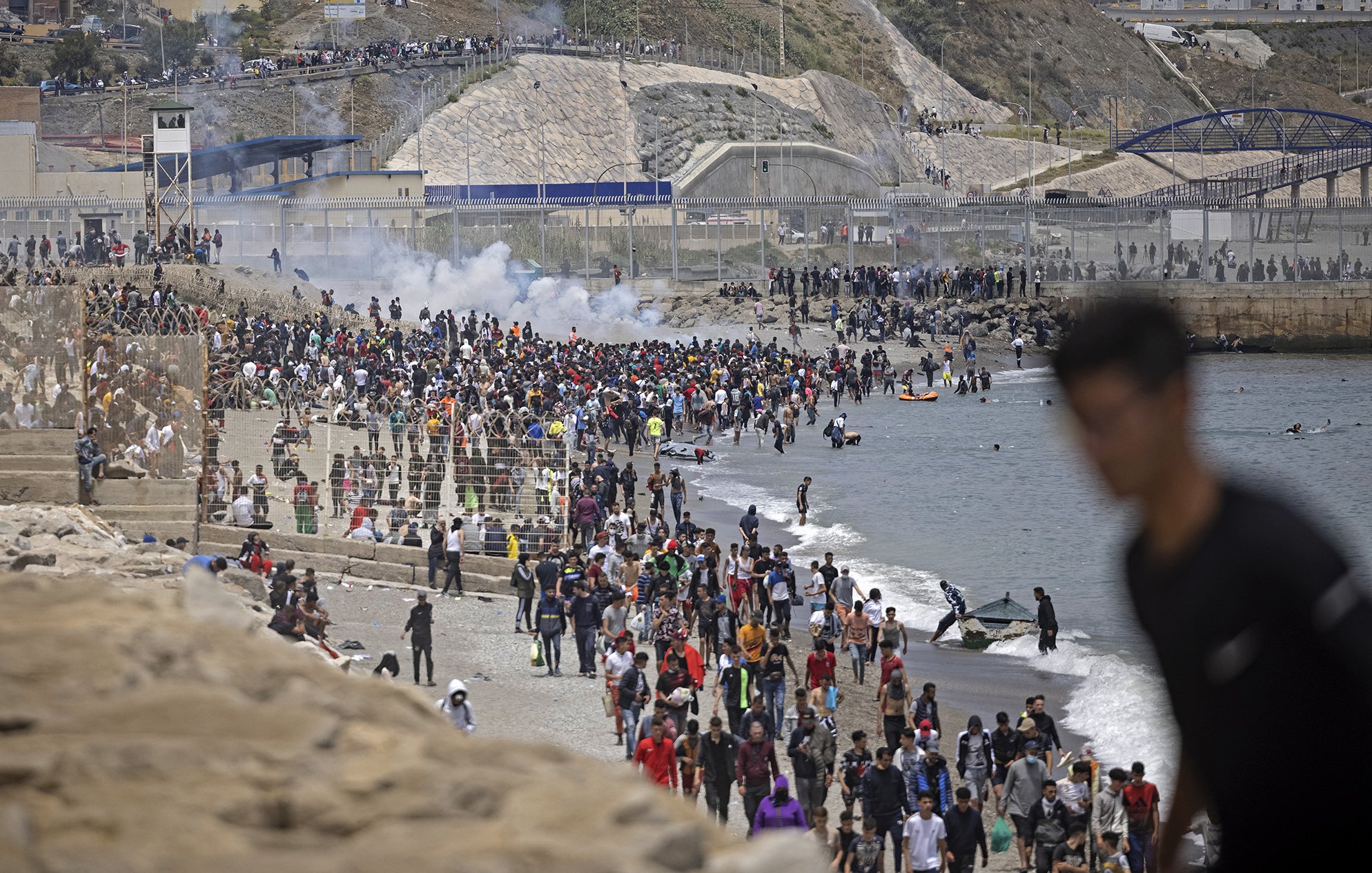 L'image montre une foule nombreuse sur une plage, avec des personnes rassemblées près de l'eau et d'autres sur le rivage. En arrière-plan, on peut voir des collines ainsi que des structures de béton. Il y a également des éléments de chaos, comme de la fumée, ce qui pourrait indiquer une situation tendue ou un événement significatif. Des gens semblent se déplacer, certains en direction de la mer, tandis que d'autres se trouvent sur le sable ou dans l'eau.