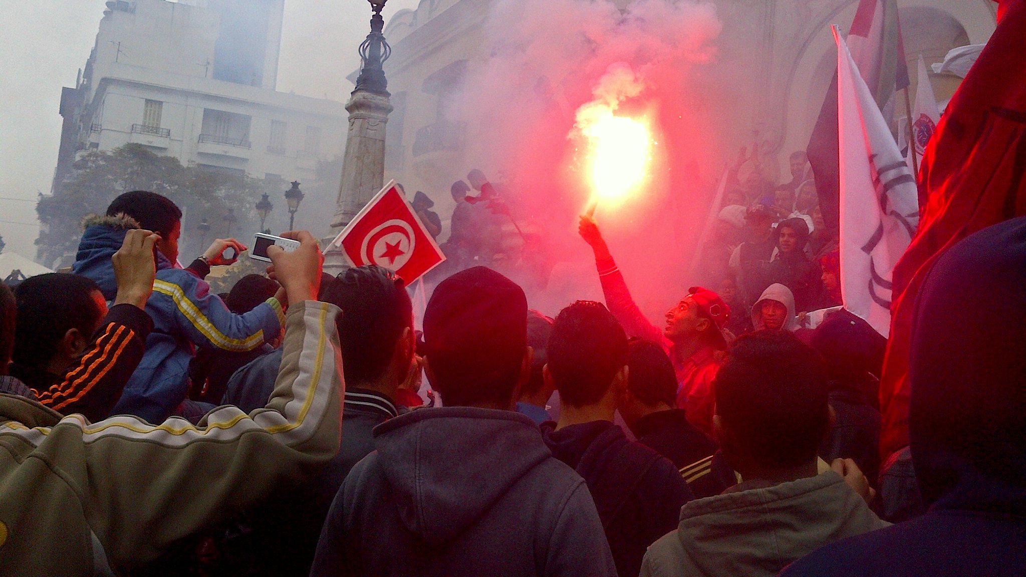 L'image montre une foule rassemblée dans une manifestation. Des personnes tiennent des drapeaux, dont un drapeau tunisien, et certains agitent des fumigènes qui dégagent de la fumée rouge. L'ambiance semble énergique et engagée, avec des bâtiments en arrière-plan. La foule paraît déterminée et unie autour de leur cause.