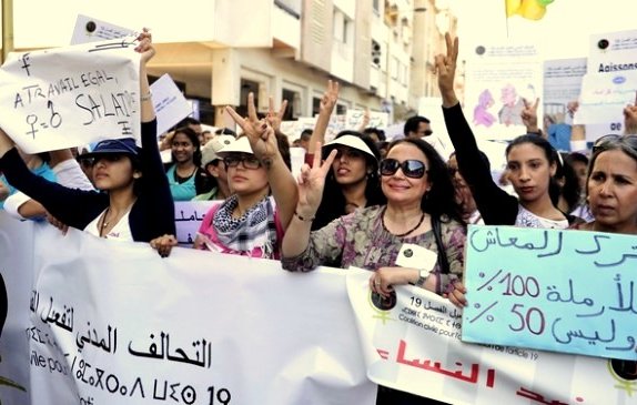 L'image montre un groupe de femmes participant à une manifestation. Elles tiennent des banderoles et des pancartes, exprimant des revendications liées aux droits des femmes. Certaines d'entre elles lèvent les mains en faisant le signe de la victoire. L'ambiance semble engagée et déterminée, avec des expressions de solidarité parmi les participantes.