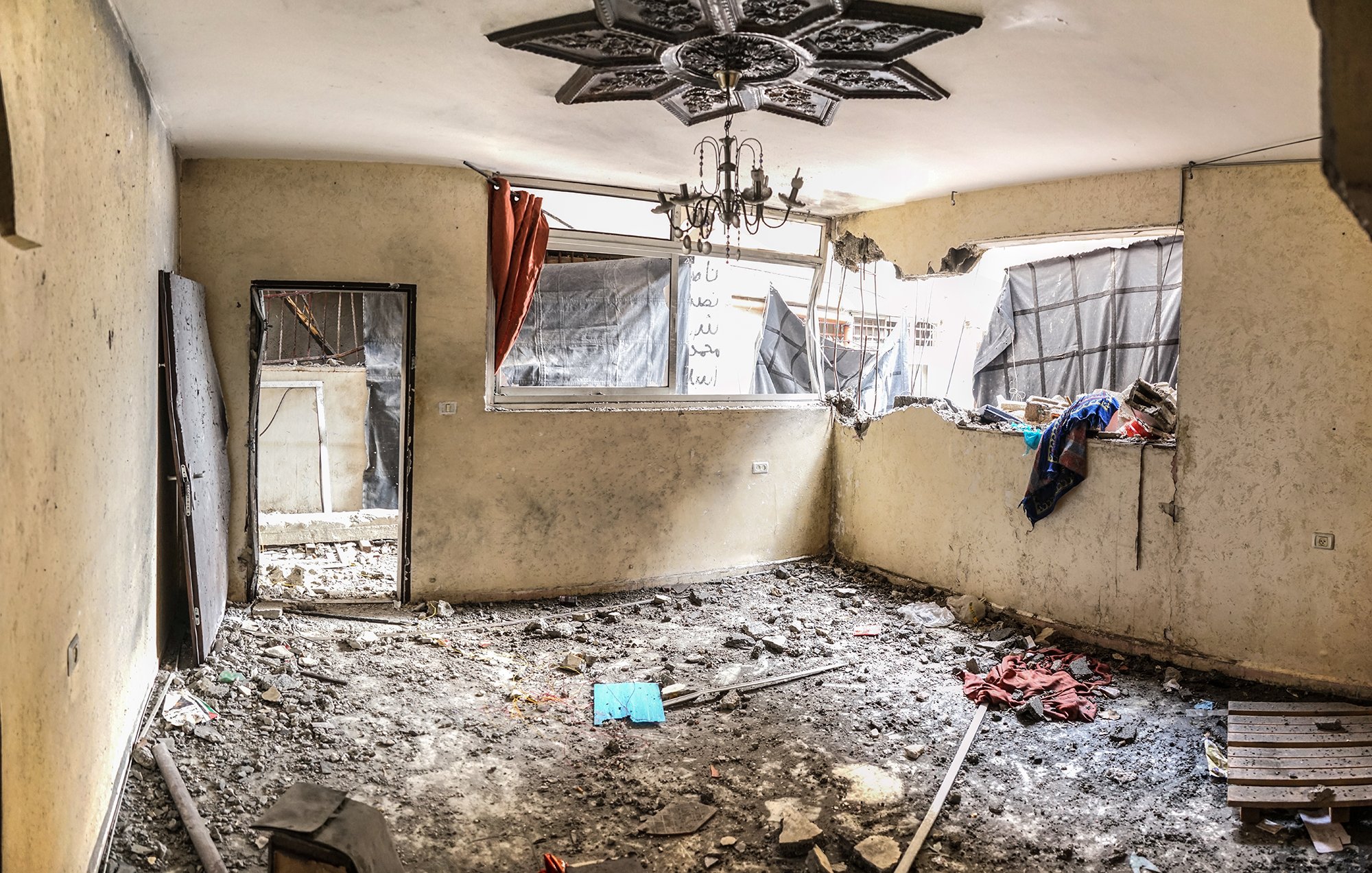 L'image montre une pièce intérieure en mauvais état. Les murs sont abîmés et la peinture est écaillée. Il y a des débris et des gravats sur le sol, ce qui indique un manque d'entretien ou des dommages récents. Une fenêtre est brisée, laissant passer la lumière et révélant des rideaux déchirés. Des vêtements et d'autres objets sont éparpillés, renforçant l'impression de désordre et d'abandon. Un lustre pend du plafond, ajoutant un contraste avec l'état délabré de l'espace. L'ambiance générale semble triste et négligée.