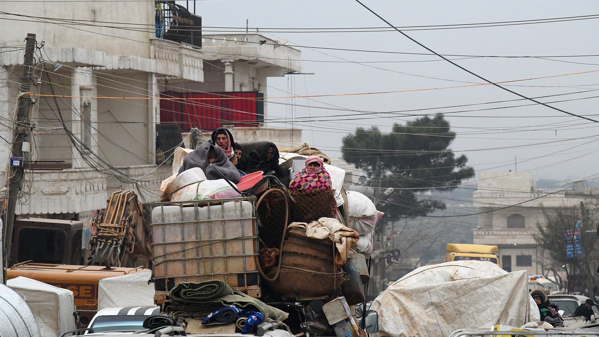 L'image montre une scène urbaine chargée, où des personnes sont perchées sur une pile de marchandises. Elles semblent se déplacer sur un véhicule surchargé, avec différents types de bagages et de matériaux. L'environnement est voilé par une lumière tamisée, peut-être due à un temps nuageux ou brumeux, et des câbles électriques traversent la scène. Les bâtiments en arrière-plan suggèrent un contexte urbain. Cette image évoque un sentiment de mouvement et de survie dans un espace urbain encombré.