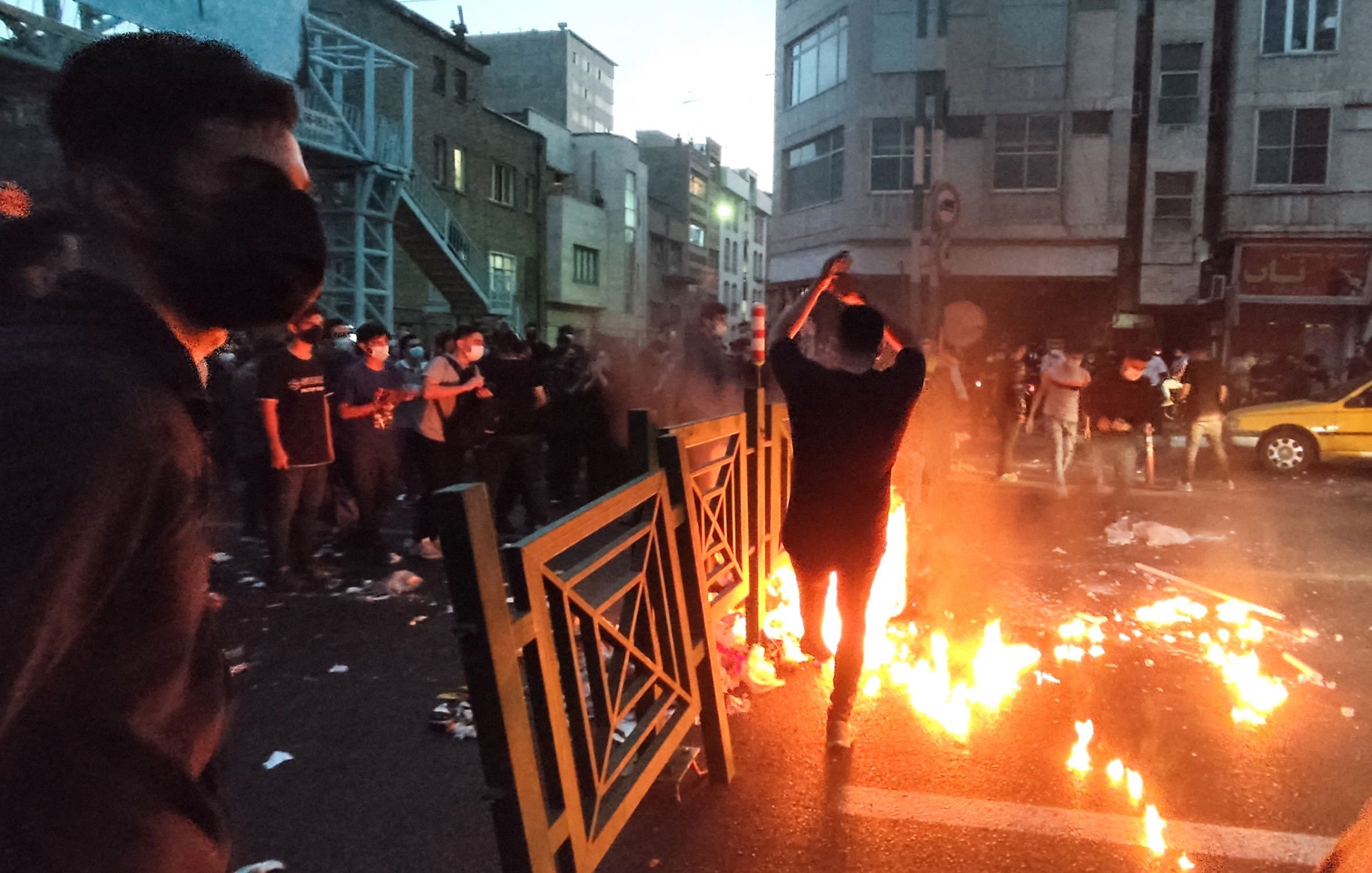 L'image montre une scène dynamique dans une rue urbaine, probablement lors d'une manifestation. On peut voir plusieurs personnes, dont l'une est au premier plan, se tenant près de flammes qui brûlent sur la route. L'atmosphère semble tendue, avec des barrières et des débris éparpillés. La lumière des flammes éclaire les visages des manifestants, qui portent des masques. En arrière-plan, on aperçoit des bâtiments urbains et une lumière tamisée, ce qui donne une impression d'urgence et de mouvement.
