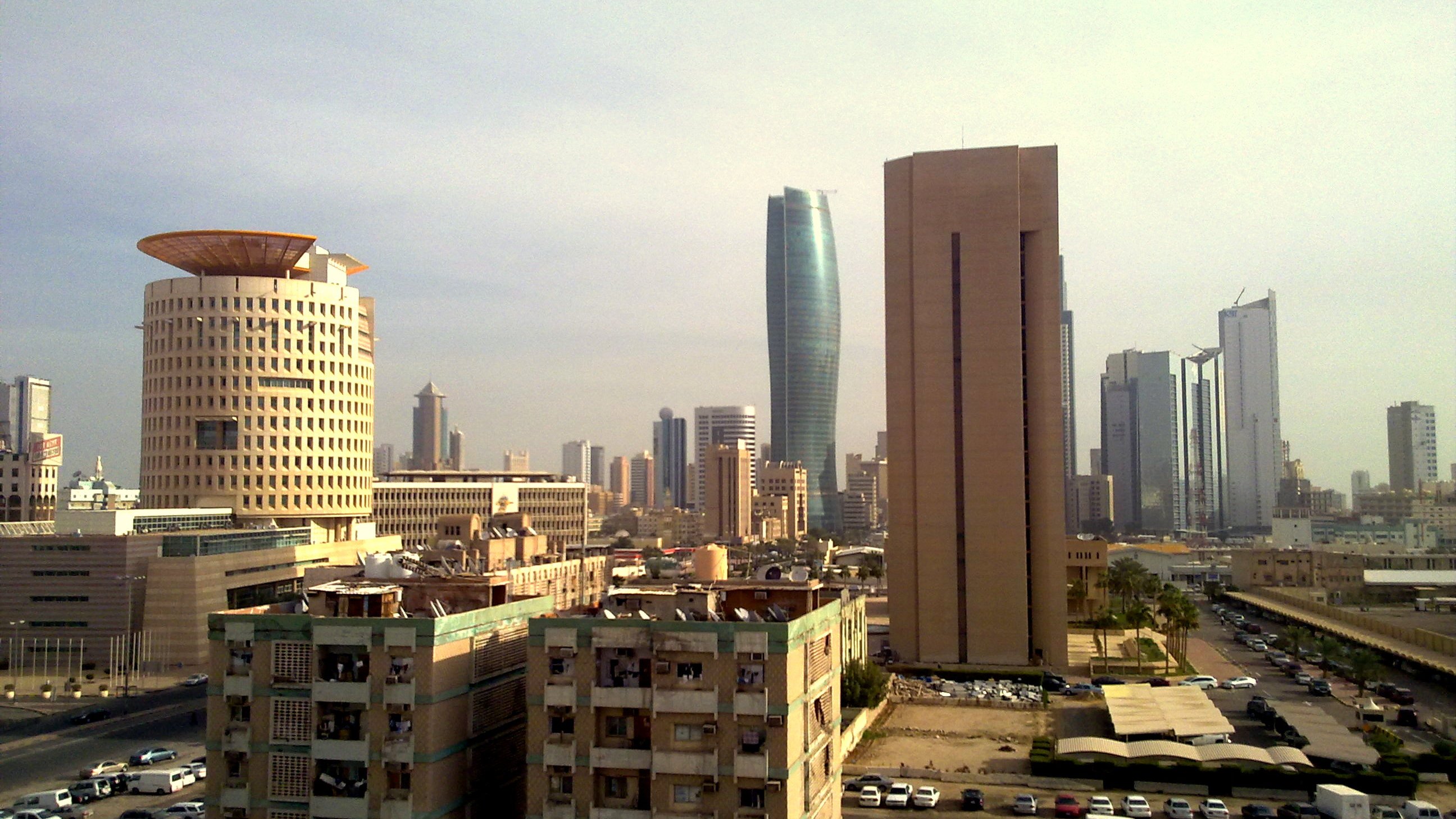 L'image montre une vue urbaine d'une grande ville moderne. On peut apercevoir plusieurs gratte-ciel aux architectures variées, avec des bâtiments à la fois anciens et contemporains. À gauche, un bâtiment au design unique, semblable à un tambour, et à droite, une structure plus élancée et moderne. En bas, on voit des immeubles vieillissants et des espaces de stationnement. Le ciel est légèrement nuageux, donnant une atmosphère calme à la scène. L'ensemble dépeint un mélange d'éléments historiques et modernes d'une métropole dynamique.