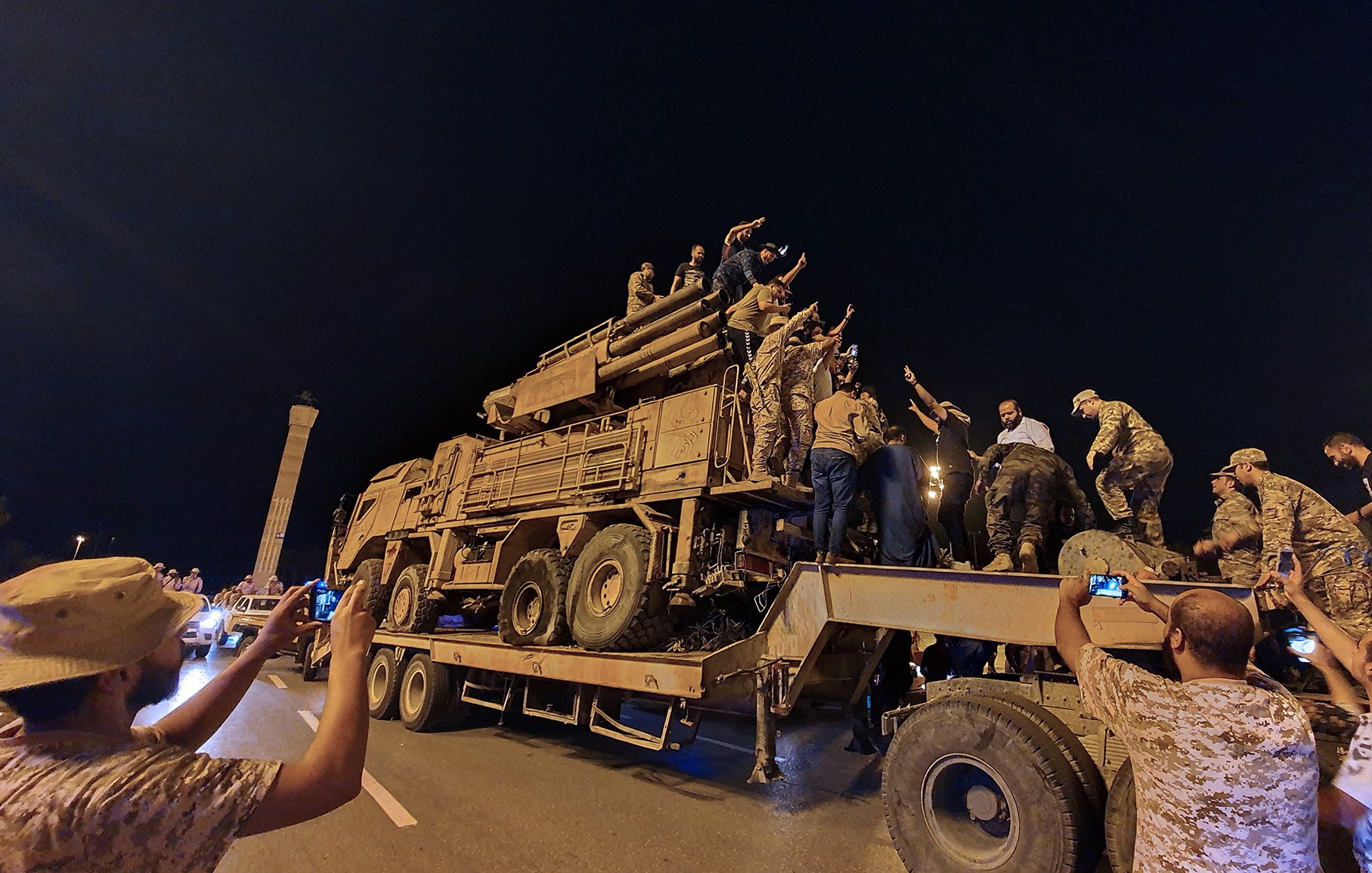 L'image montre un groupe de personnes rassemblées autour d'un véhicule militaire sur une route, probablement pendant la nuit. Plusieurs individus sont montés sur le véhicule et semblent exprimer des émotions fortes, peut-être en célébrant ou en manifestant un soutien. D'autres personnes prennent des photos avec leurs téléphones. L'ambiance générale semble dynamique et impliquée, avec une forte présence militaire sur le site.