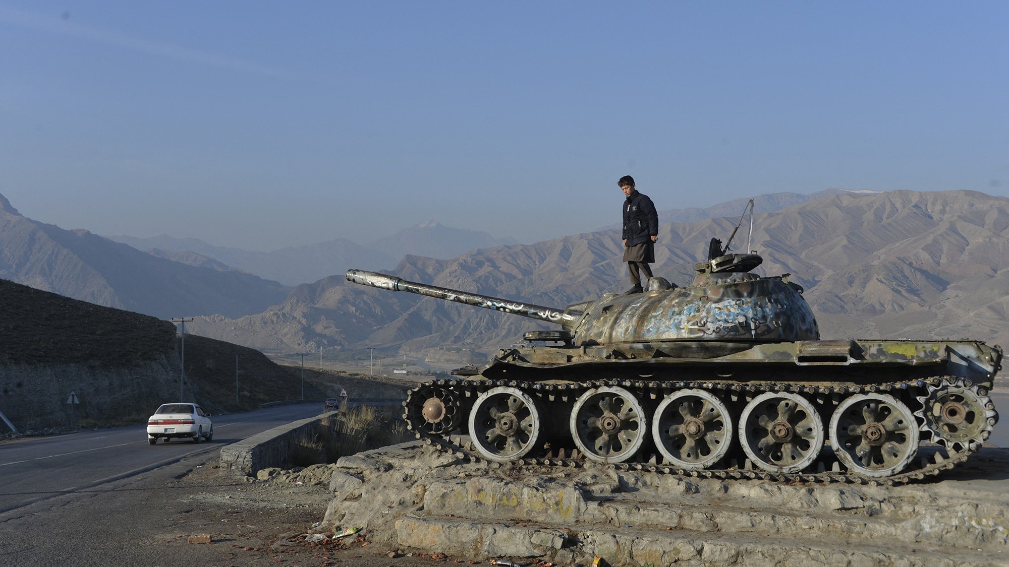 L'image montre un homme debout sur un char de bataille, probablement un ancien char T-55, qui est posé sur une plateforme. Le char présente des signes d'usure et de rouille, et le paysage environnant est montagneux avec un ciel dégagé. On peut voir une route à proximité, ainsi qu'une voiture blanche circulant. L'ambiance évoque un mélange de nostalgie et de désolation, rappelant une époque passée de conflits.