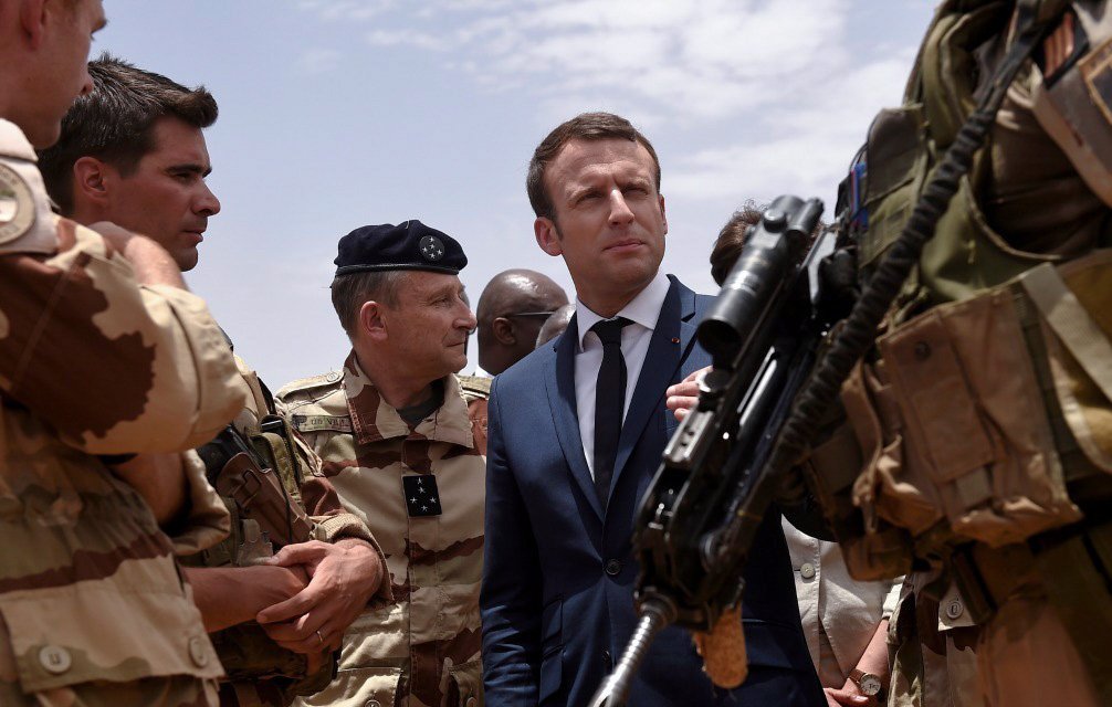 L'image montre un groupe de militaires en uniforme, dont certains portent des casques et des armes, rassemblés autour d'un homme en costume et cravate. Cet homme a une expression concentrée et semble en conversation avec les militaires, indiquant une interaction sérieuse. Le décor évoque un environnement militaire, probablement en extérieur, sous un ciel partiellement nuageux. Cette scène pourrait représenter une visite officielle ou une interaction avec des forces armées.