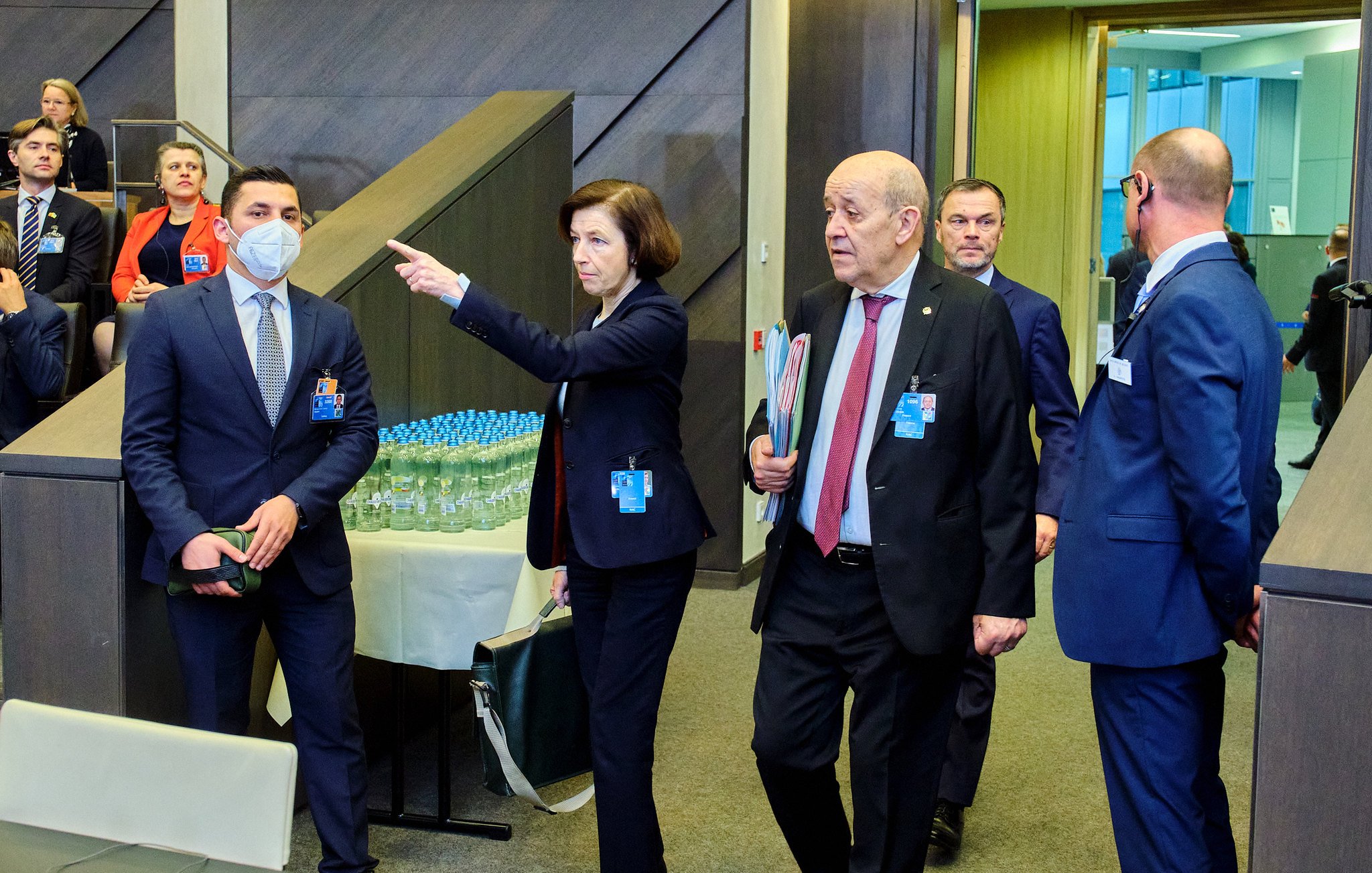 L'image montre un groupe de personnes dans un environnement de conférence ou de réunion. Au centre, une femme portant un blazer indique quelque chose, tandis que d'autres personnes, dont un homme en costume et en cravate, l'écoutent attentivement. Certains participants semblent en discussion, tandis que d'autres regardent dans la direction où la femme pointe. En arrière-plan, on peut voir des bouteilles d'eau sur une table. L'atmosphère semble sérieuse et professionnelle.