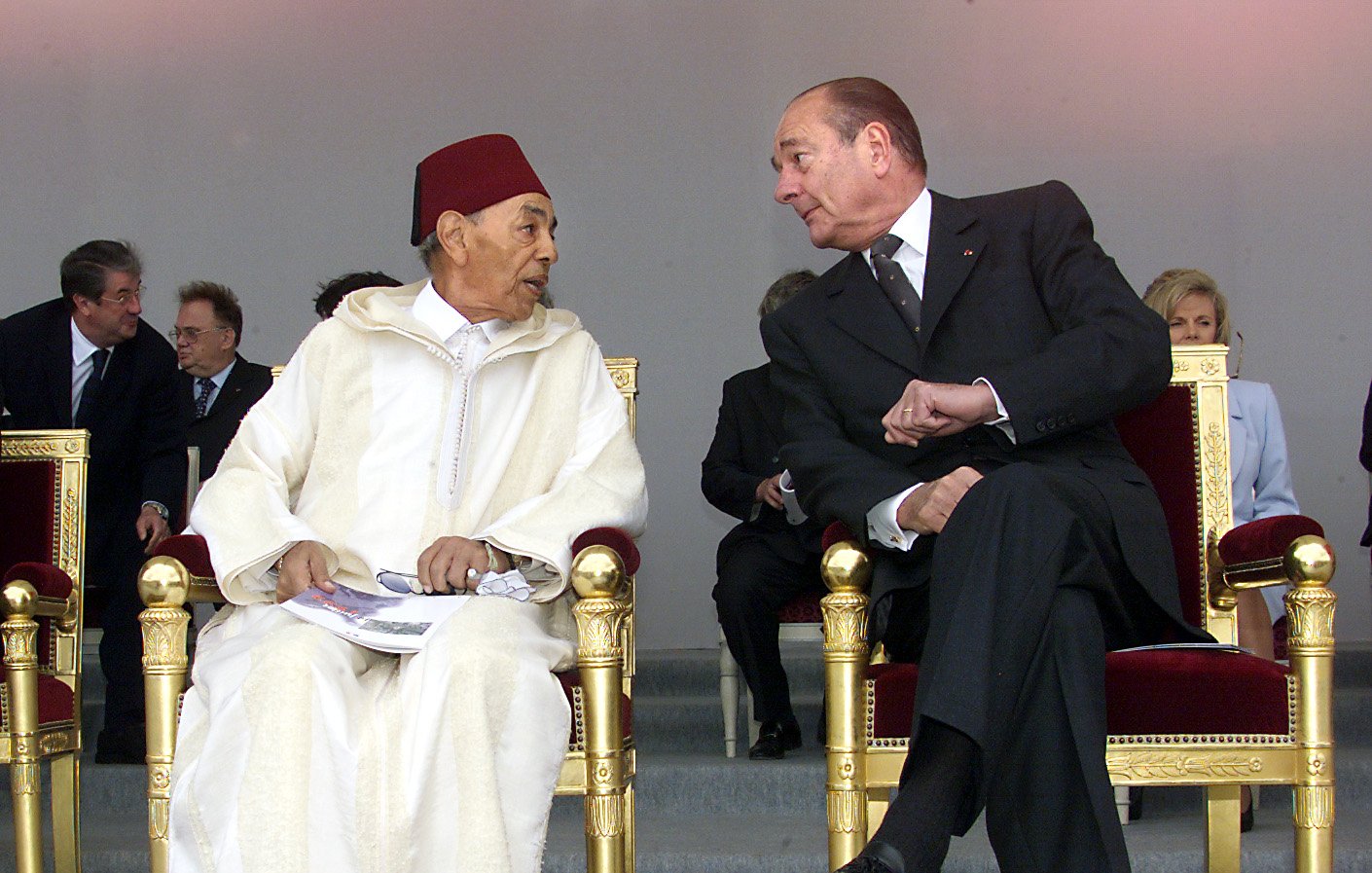 L'image montre deux hommes assis sur des chaises ornées, dans un cadre formel. L'un porte un habit traditionnel marocain avec un fez rouge, tandis que l'autre est vêtu d'un costume noir. Ils semblent engagés dans une conversation, se regardant avec attention. En arrière-plan, on aperçoit d'autres personnes, indiquant que cette scène a lieu lors d'un événement officiel. Leurs expressions semblent amicales et concentrées.