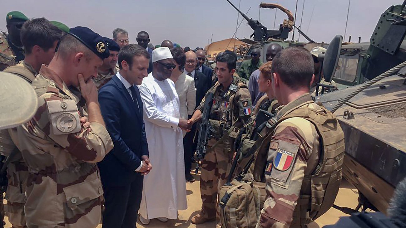 L'image montre un groupe de militaires et de responsables, probablement en situation de rencontre officielle dans un environnement désertique. Au centre, on peut voir un homme en costume, représentant vraisemblablement un leader, en train de discuter avec des soldats équipés de matériel militaire. Les militaires sont en uniforme et semblent engagés dans une mission, tandis que d'autres personnes obéissent à la dynamique de la réunion. L'atmosphère paraît sérieuse et solennelle.