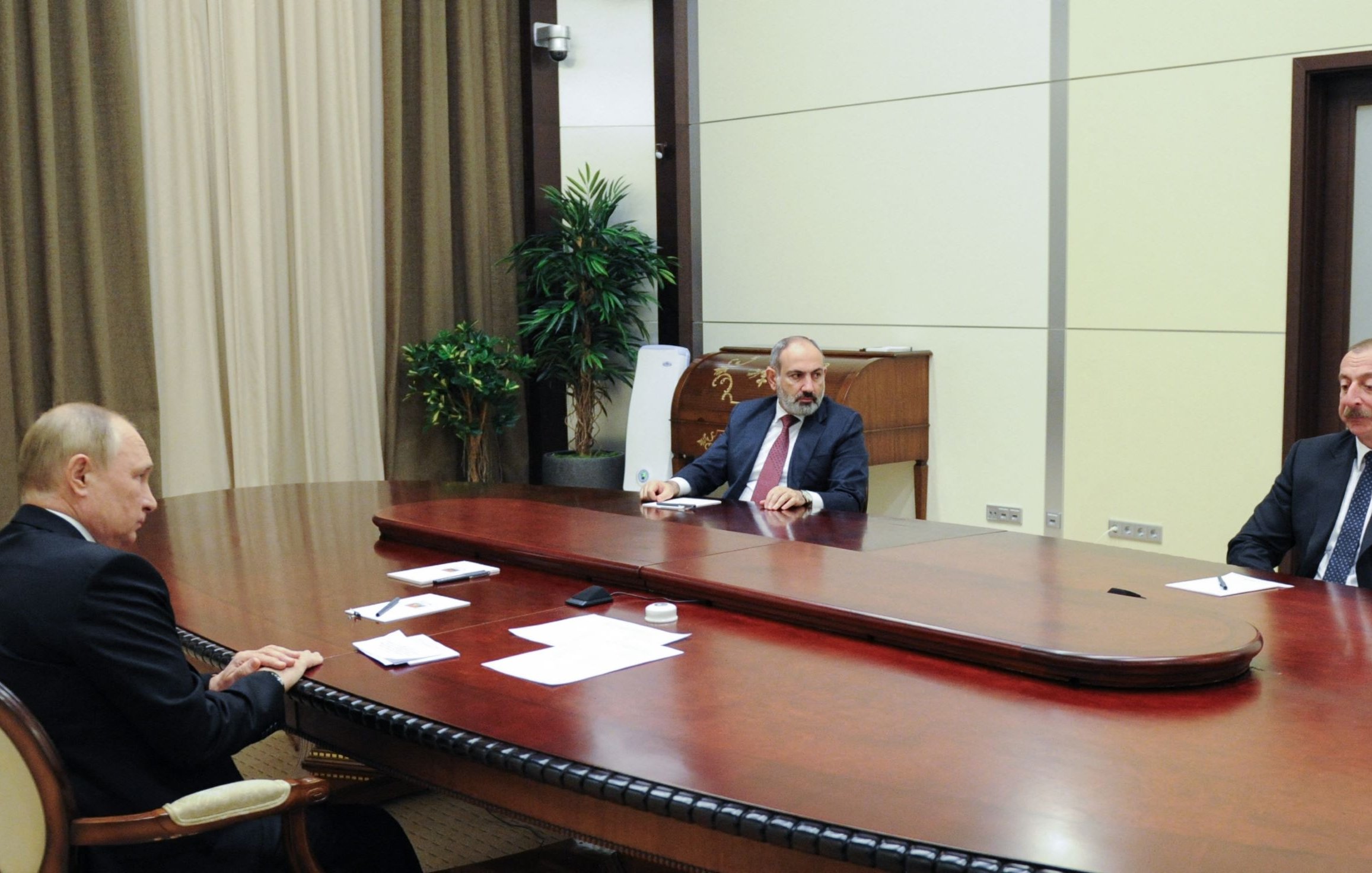 L'image montre une réunion formelle dans une salle ornée. Trois hommes sont assis autour d'une grande table ovale en bois. Au centre, un homme en costume parlant, probablement un chef d'État ou un dirigeant. À sa gauche, un homme au visage sérieux, également en costume. À droite, un autre homme, avec une expression attentive. La pièce est décorée de rideaux et d'une plante d'intérieur, ce qui lui donne un aspect sobre et professionnel.