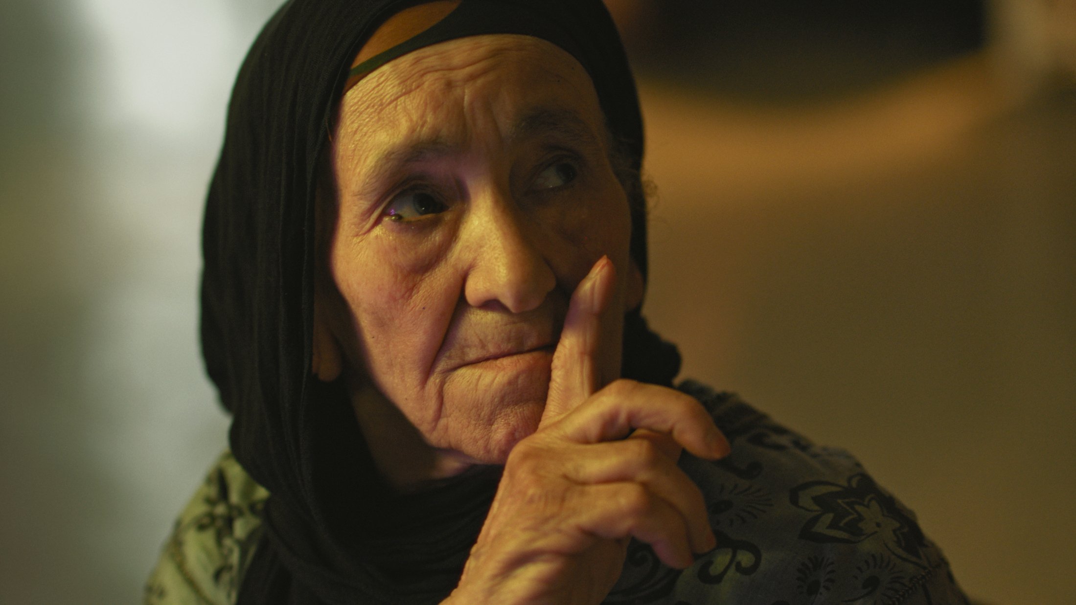 L'image montre une femme âgée avec un regard pensif. Elle porte un foulard noir qui encadre son visage. Son expression est sérieuse et elle semble faire un geste de silence avec son doigt posé sur ses lèvres. La lumière dans le fond est douce, créant une ambiance calme et introspective. Les détails de son visage reflètent une certaine sagesse et vécu.