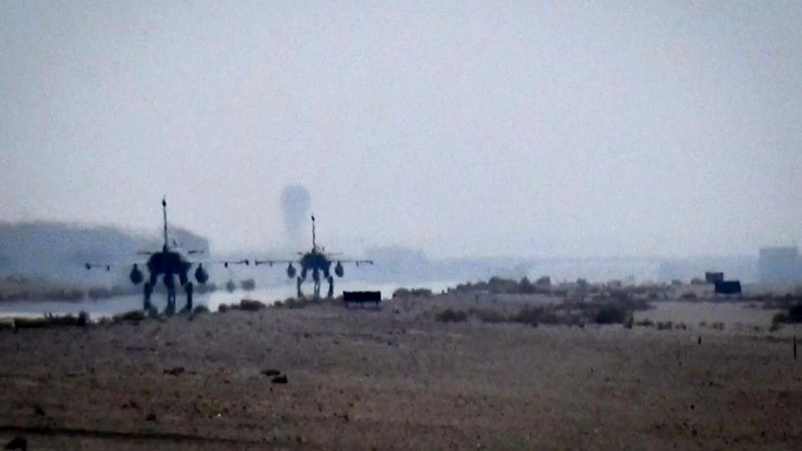 L'image montre deux avions militaires aperçus au loin dans un paysage désertique. L'atmosphère est légèrement brumeuse, ce qui donne une impression de profondeur. On peut distinguer une piste d'atterrissage ou d'envol, et le sol semble aride avec peu de végétation. Les avions sont visibles, mais les détails sont flous en raison de la distance et de la qualité de la prise de vue.