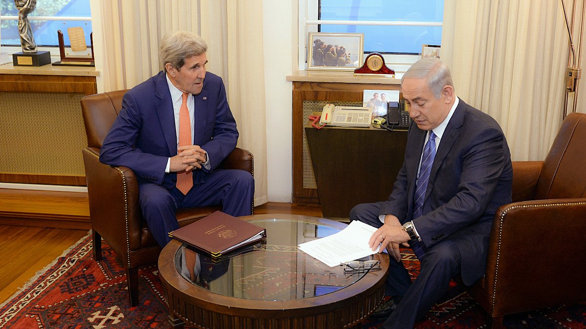 L'image montre deux hommes assis dans une pièce restructurée. L'un est vêtu d'un costume orange et l'autre d'un costume sombre, tous deux semblent être en discussion sérieuse. Ils sont assis autour d'une table basse en bois, avec des documents devant eux. En arrière-plan, on aperçoit une fenêtre et une décoration sobre, suggérant un cadre officiel et professionnel.