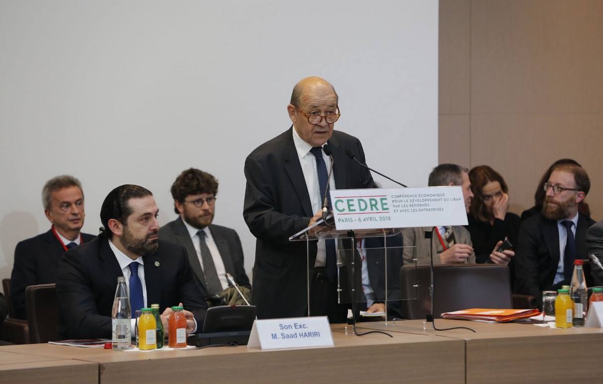 L'image montre un homme âgé, probablement un orateur, se tenant derrière un podium avec un panneau portant le mot "CEDRE" et la date de l'événement. Il semble prendre la parole lors d'une conférence. Autour de lui, plusieurs autres personnes sont assises, attentives, et il y a des bouteilles d'eau et des boissons sur la table. L'ambiance semble formelle et professionnelle, suggérant un événement important.