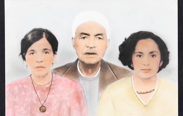 L'image représente un portrait de famille en couleur, où l'on voit trois personnes. Au centre se trouve un homme âgé avec une barbe grise, portant un chapeau blanc et un vêtement sobre. De chaque côté de lui, il y a deux femmes. À gauche, une femme avec des cheveux sombres, portant un haut rose et un collier. À droite, une autre femme avec des cheveux bouclés, habillée d'un haut jaune, qui affiche également un collier. L'arrière-plan est lumineux et flou, mettant en valeur les visages des sujets.