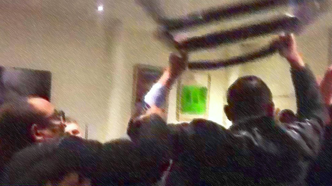 L'image montre un groupe de personnes se rassemblant autour d'une bannière ou d'un drapeau, qu'ils soulèvent. L'atmosphère semble dynamique et engagée, avec des individus exprimant leurs émotions. Le fond est flou, ce qui met l'accent sur l'action au premier plan. On peut sentir une ambiance de solidarité ou de protestation.