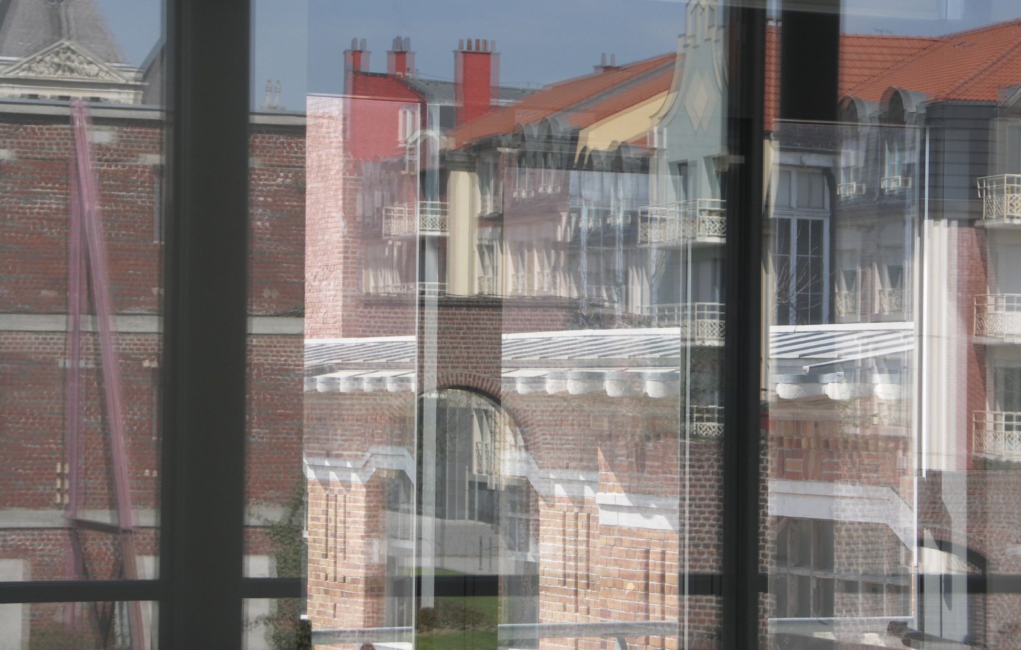 L'image montre un jeu de reflets à travers de grandes fenêtres. On peut apercevoir des bâtiments aux façades en briques et des toits de différentes couleurs. Les reflets créent une composition visuelle intrigante, mêlant les éléments extérieurs et la transparence des vitres. La lumière naturelle accentue les détails des structures architecturales.
