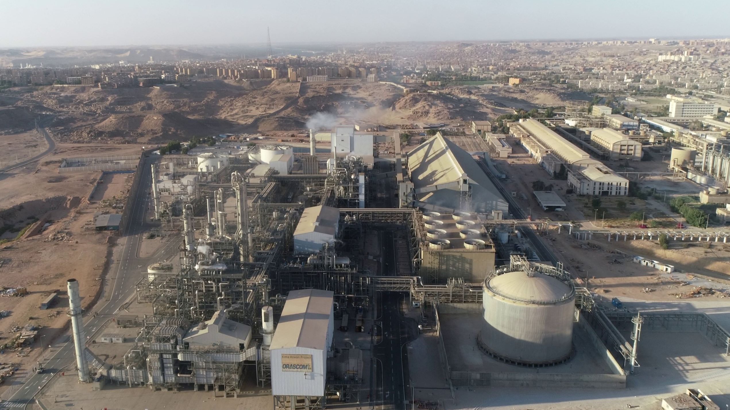 L'image présente une vue aérienne d'un complexe industriel. On peut observer plusieurs bâtiments de grande taille, comprenant des structures cylindriques et pyramidales. La zone environnante semble aride avec des collines et des bâtiments à l'arrière-plan. Des tuyaux et des infrastructures industrielles sont visibles, indiquant une activité de production. On discerne également de la vapeur s'échappant d'une des installations, suggérant un processus en cours. L'ensemble dénote un environnement industriel dans une région désertique.