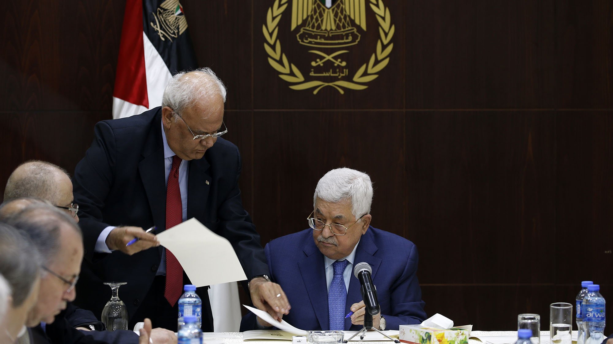 L'image montre une réunion, probablement politique, avec plusieurs hommes assis à une table. Au premier plan, un homme, probablement un leader ou un officiel, est assis regardant des documents. Un autre homme, debout à ses côtés, semble lui présenter des papiers ou des informations. En arrière-plan, on peut voir un drapeau, ainsi qu'un emblème qui semble représenter une organisation ou un gouvernement. L'ambiance générale de la scène est sérieuse et attentive, suggérant une discussion importante.