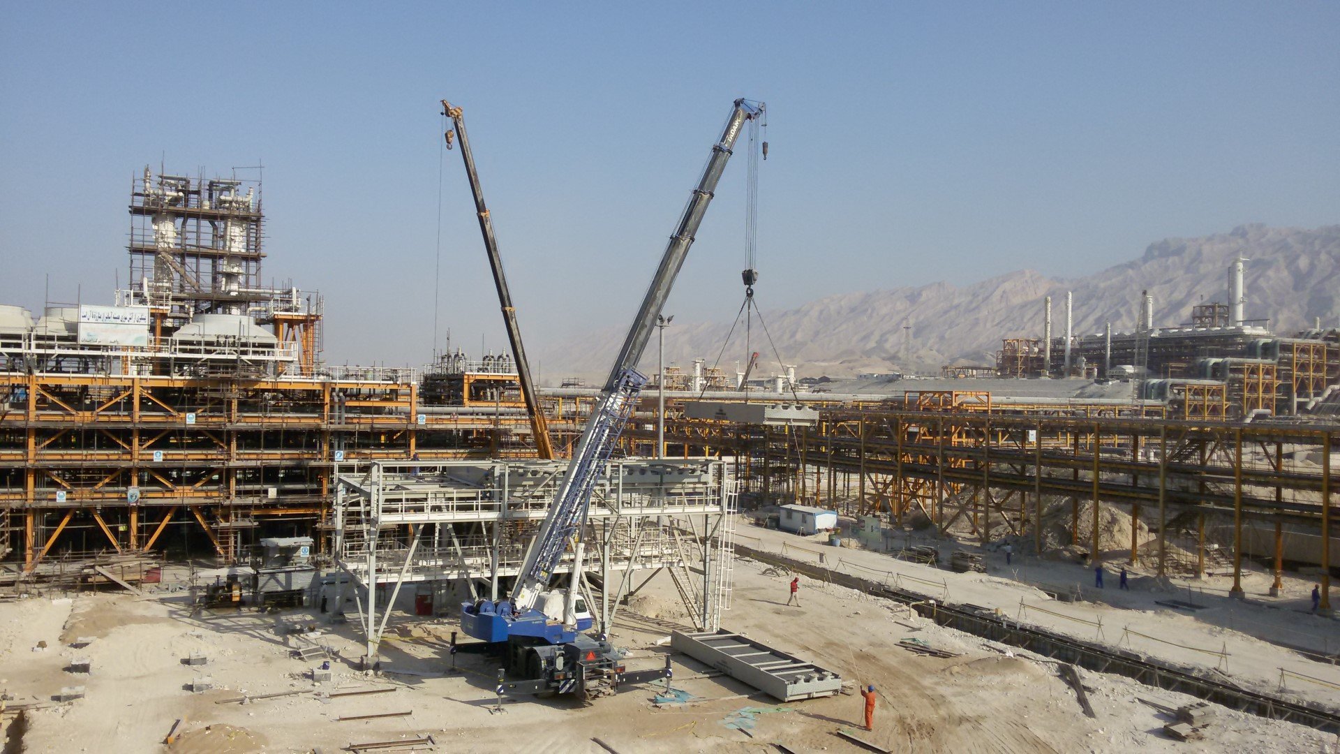 L'image montre un chantier de construction avec plusieurs grues sur le site. On peut voir des structures métalliques en voie d'achèvement à l'arrière-plan, ainsi que des véhicules de chantier et des ouvriers sur le site. Le paysage environnant est montagneux, et le ciel est dégagé, ce qui suggère une journée ensoleillée. Le chantier semble être à un stade de construction avancé, avec des matériaux disposés sur le sol.