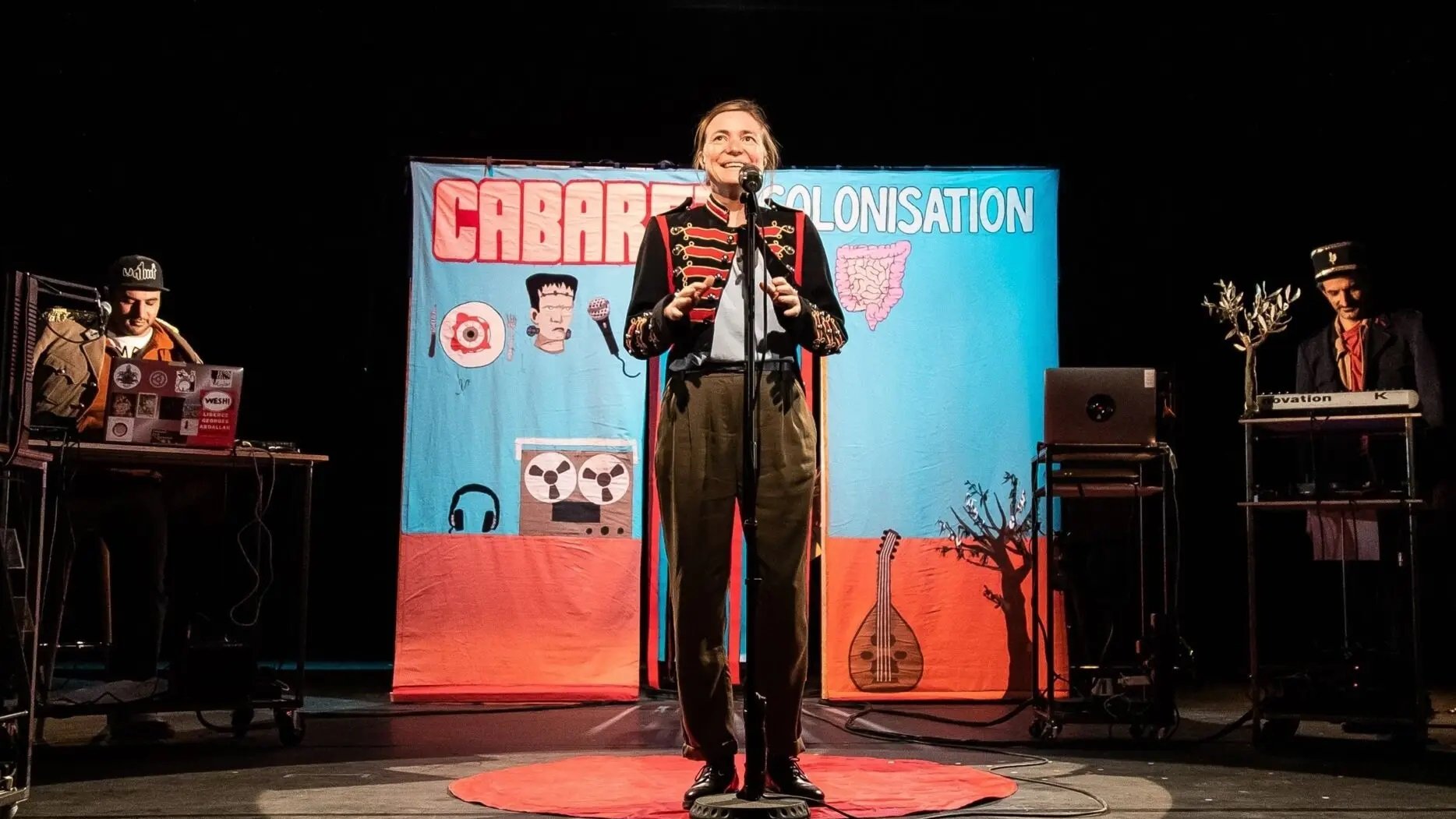 L'image montre une scène de spectacle, probablement un cabaret. Au centre, un artiste souriant est sur scène avec un micro, habillé de manière théâtrale. En arrière-plan, un grand panneau coloré affiche le mot "CABARET" et "COLONISATION", suggérant un thème spécifique. De chaque côté, on aperçoit des musiciens ou des techniciens avec des instruments ou des équipements, contribuant à l'atmosphère festive et engageante. Le sol est orné d'un tapis rouge, ajoutant une touche de mise en scène au spectacle.