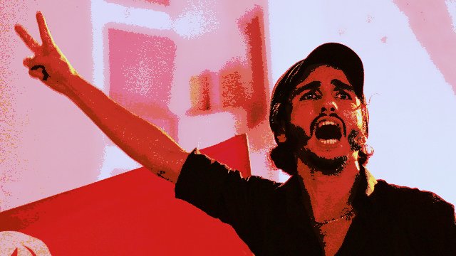 L'image montre un homme debout, apparemment engagé dans une expression de passion ou de colère. Il lève son bras en faisant un signe de victoire. Son visage est très expressif, avec une bouche ouverte comme s'il était en train de crier ou de s'exprimer avec force. En arrière-plan, on peut discerner un drapeau, qui pourrait suggérer un contexte politique ou social. Les couleurs de l'image sont assez vives, ce qui accentue l'intensité de la scène.