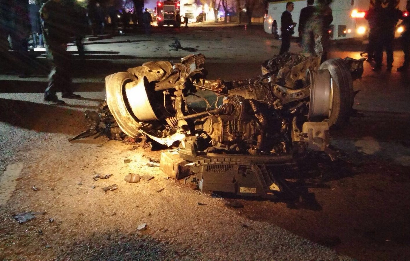 L'image montre un véhicule gravement endommagé, probablement retourné sur le côté. On peut voir des débris éparpillés sur la route, indiquant un accident violent. La scène est éclairée par des lumières, suggérant la présence de services d'urgence ou de témoins. Le contexte semble être nocturne, ce qui accentue les dégâts visibles sur le véhicule.