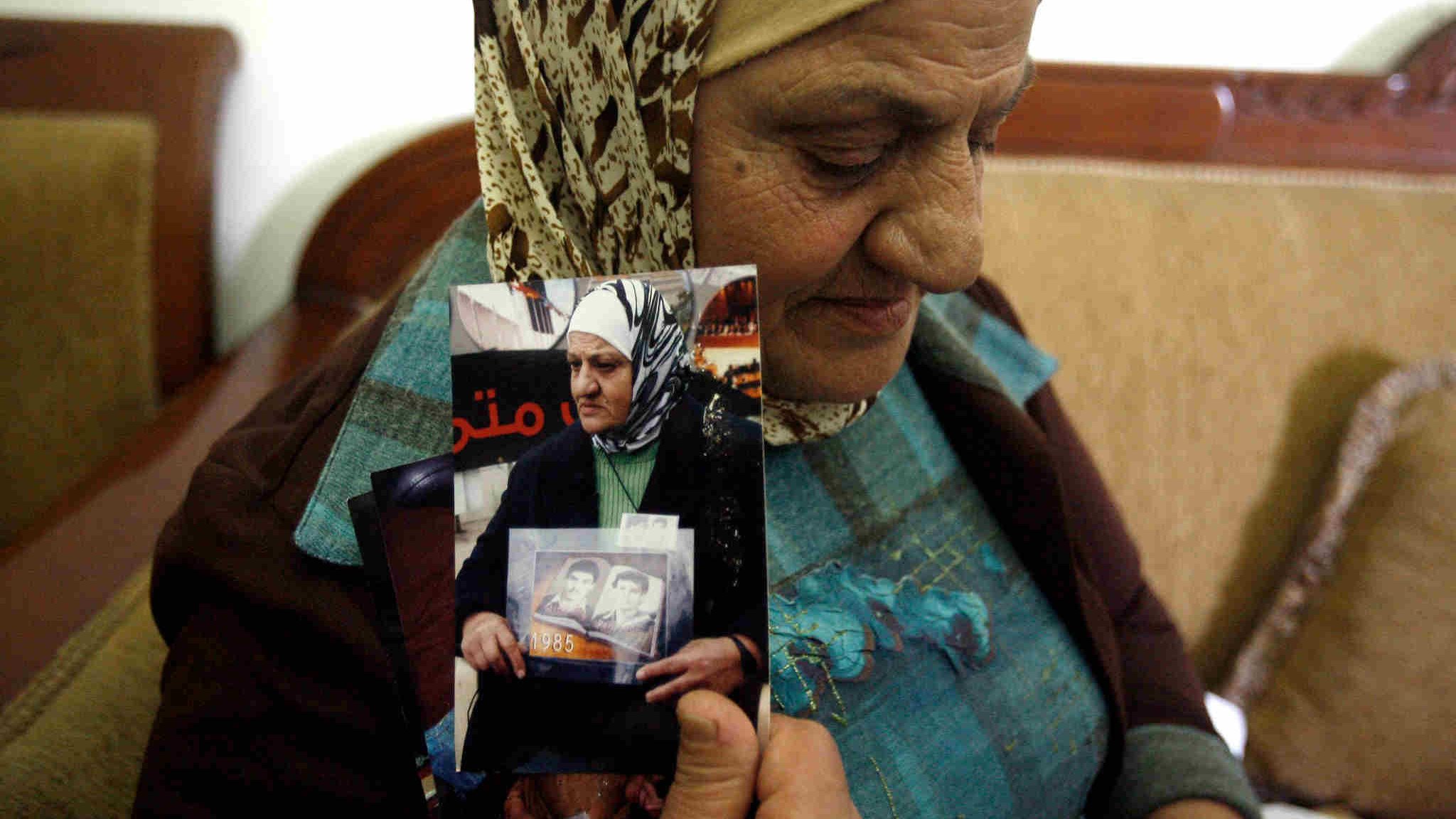 L'image montre une femme âgée portant un foulard, assise sur un canapé. Elle tient des photos en noir et blanc dans ses mains. Son expression semble empreinte de nostalgie ou de tristesse, indiquant peut-être qu'elle évoque des souvenirs douloureux. L'environnement semble simple, avec des murs clairs et un décor sobre en arrière-plan. La scène transmet une forte charge émotionnelle, suggérant un lien personnel avec les images qu'elle tient.