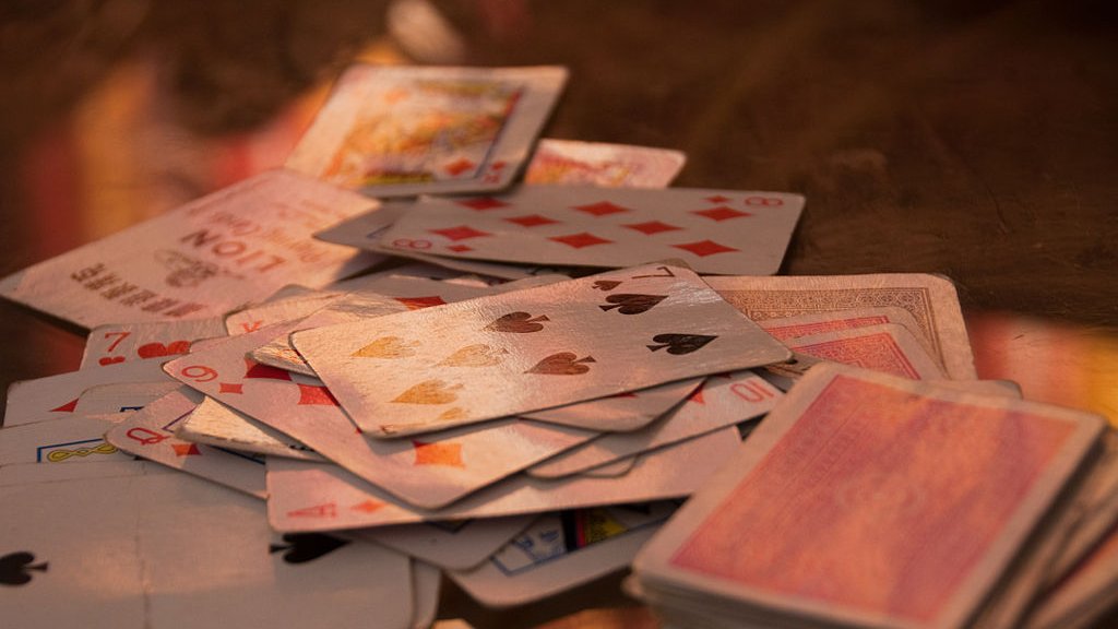 L'image montre un tas de cartes à jouer éparpillées sur une surface. On peut voir différents types de cartes, y compris des cartes de cœur, de carreau, de trèfle et de pique. Certaines cartes semblent retournées, tandis que d'autres sont face visible, montrant leurs motifs et valeurs. L'éclairage apporte une ambiance chaleureuse et met en valeur la texture des cartes.