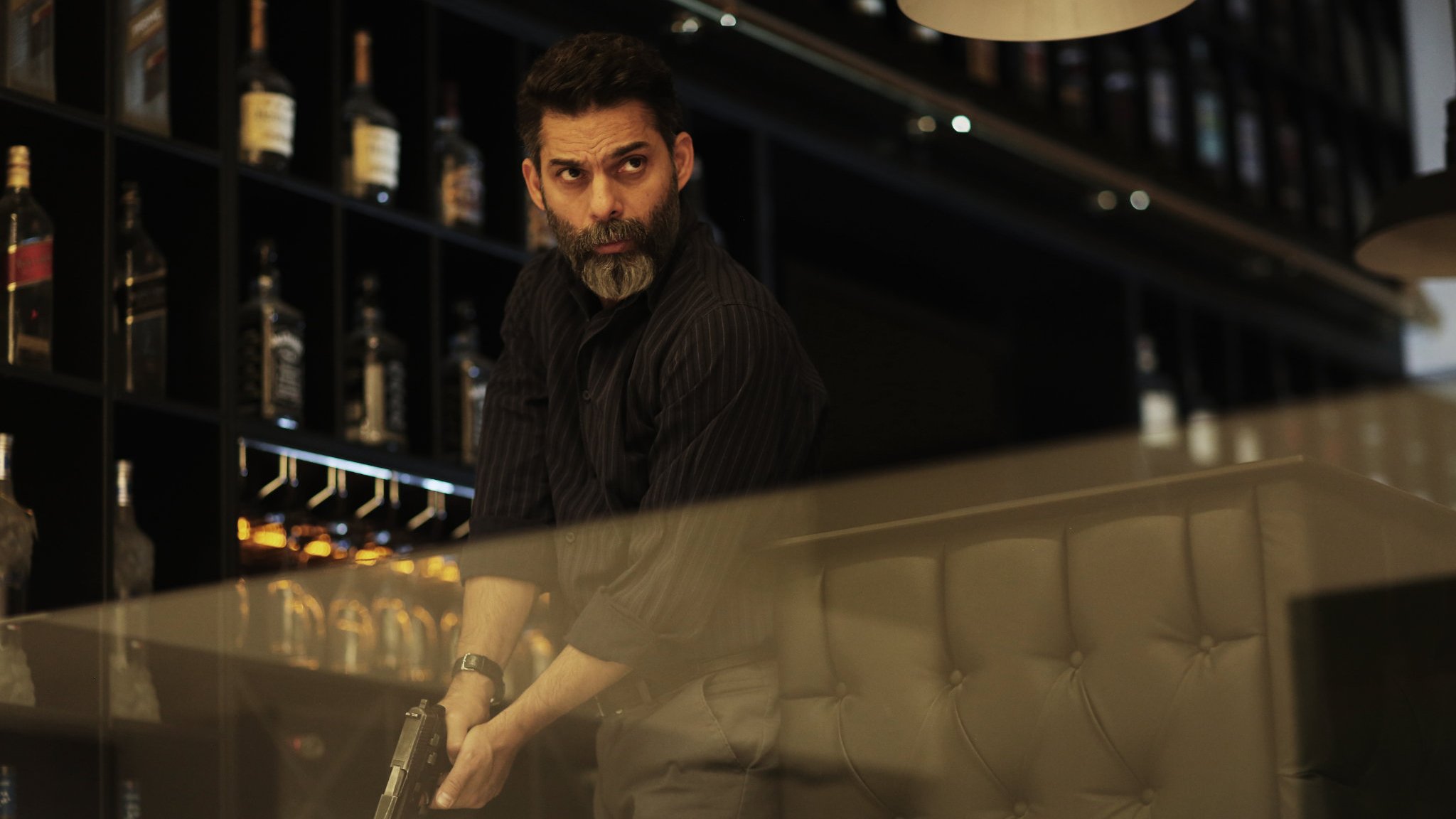 L'image montre un homme, probablement dans un bar ou un restaurant, tenant une arme à feu. Il a une barbe grise et une expression sérieuse, suggérant qu'il est dans une situation tendue. Le décor en arrière-plan comprend des étagères avec des bouteilles de spiritueux. L'éclairage est tamisé, créant une atmosphère dramatique.