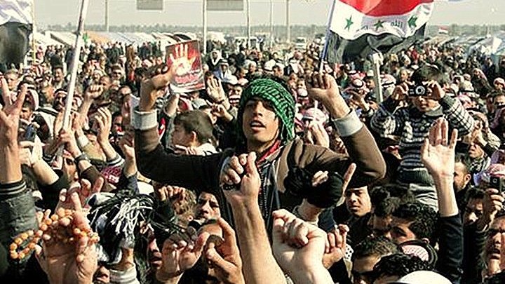 L'image montre une grande foule rassemblée, probablement lors d'une manifestation ou d'un événement public. Les personnes levées les bras, brandissant des drapeaux, affichent des expressions d'énergie et d'engagement. Certains portent des vêtements traditionnels, et l'ambiance semble dynamique et fervente. Des pancartes ou des symboles visibles pourraient suggérer un message politique ou social. L'environnement semble être un espace ouvert, peut-être une place ou une route.