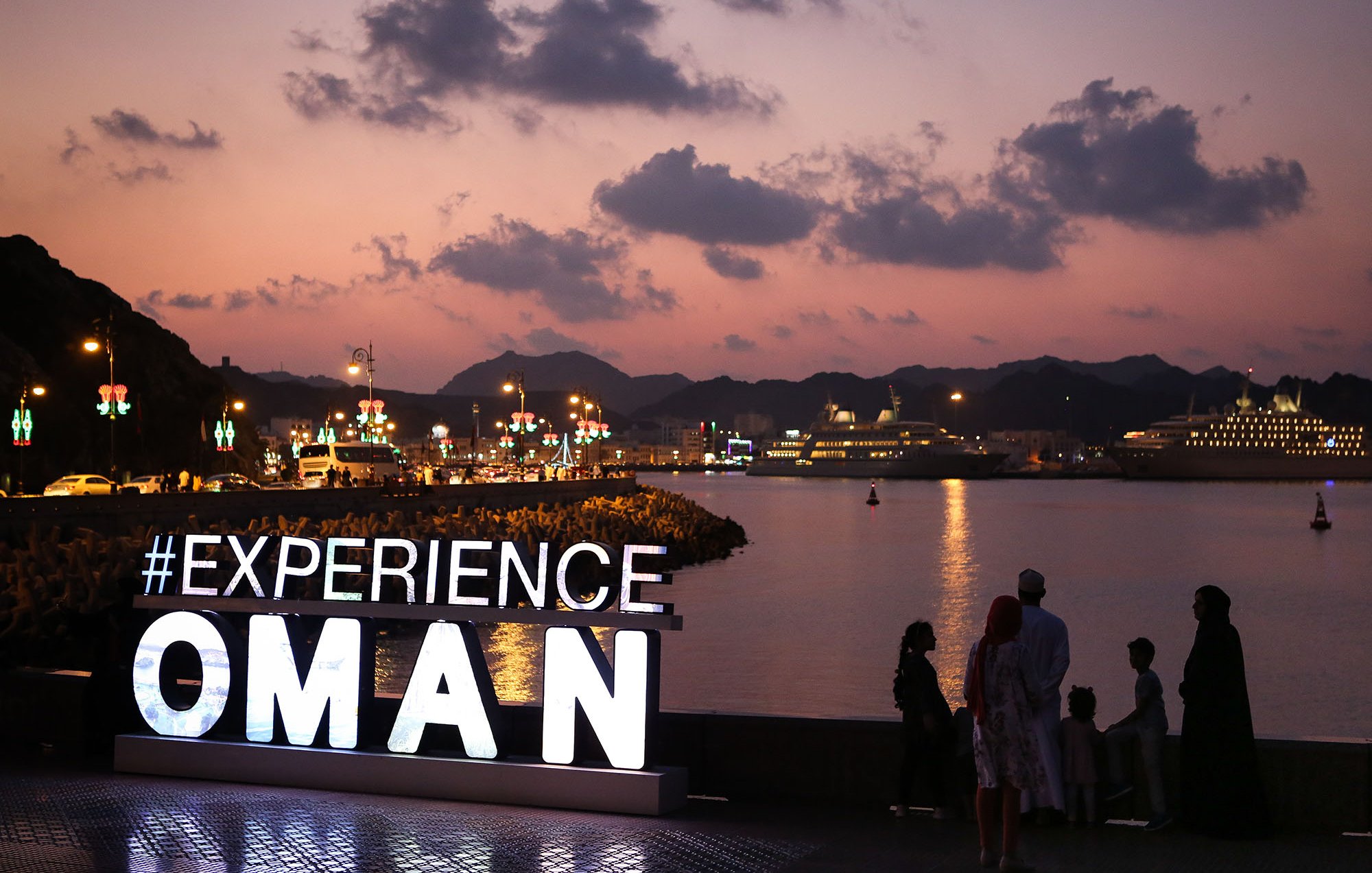 L'image montre une scène au coucher du soleil à Oman, avec des montagnes en arrière-plan et un port où se trouvent des bateaux et des navires de croisière. Au premier plan, il y a un grand panneau lumineux affichant le mot "EXPERIENCE" suivi d'"OMAN". Des personnes se tiennent devant le panneau, admirant la vue sur l'eau. La lumière douce du crépuscule et l'ambiance paisible de la scène créent une atmosphère accueillante.
