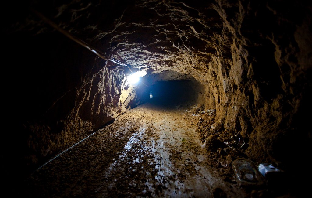 Cette image montre un tunnel souterrain. Les parois sont irrégulières et terreuses, indiquant qu'il a été creusé à la main ou à l'aide d'outils simples. Le sol est recouvert de terre, et une source de lumière artificielle éclaire l'intérieur, créant un contraste entre l'obscurité du tunnel et la lumière. À droite, on peut apercevoir des déchets éparpillés, ce qui suggère que l'endroit a été utilisé pour un certain temps. Le tunnel semble relativement étroit et évoque une atmosphère de mystère ou d'aventure.