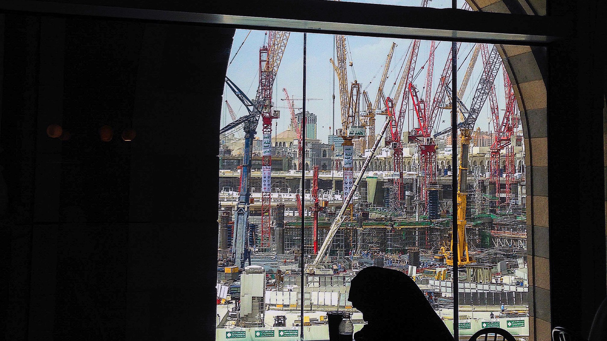 L'image montre un chantier de construction vu à travers une grande fenêtre à arc. À l'intérieur, on aperçoit une silhouette assise à une table. De l'autre côté de la vitre, de nombreux grues et engins de chantier se dressent, travaillant à un projet de grande envergure. Le ciel est partiellement dégagé, ajoutant une touche de lumière à la scène. L'ensemble dégage une atmosphère de travail intensif et de développement urbain.
